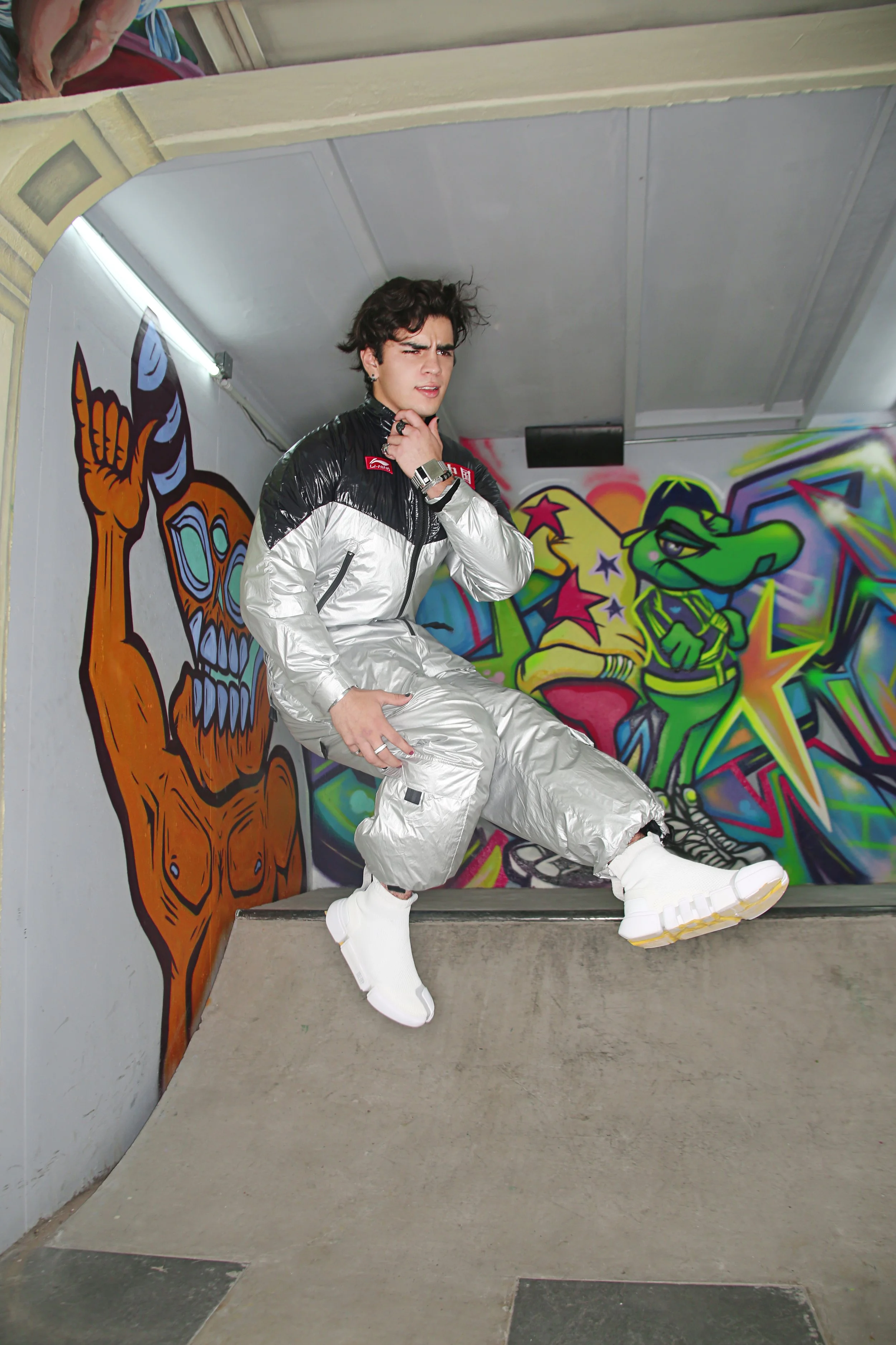 A young man with dark, curly hair poses in front of vibrant graffiti wall art, wearing a shiny silver and black jacket with matching baggy pants and white chunky sneakers.