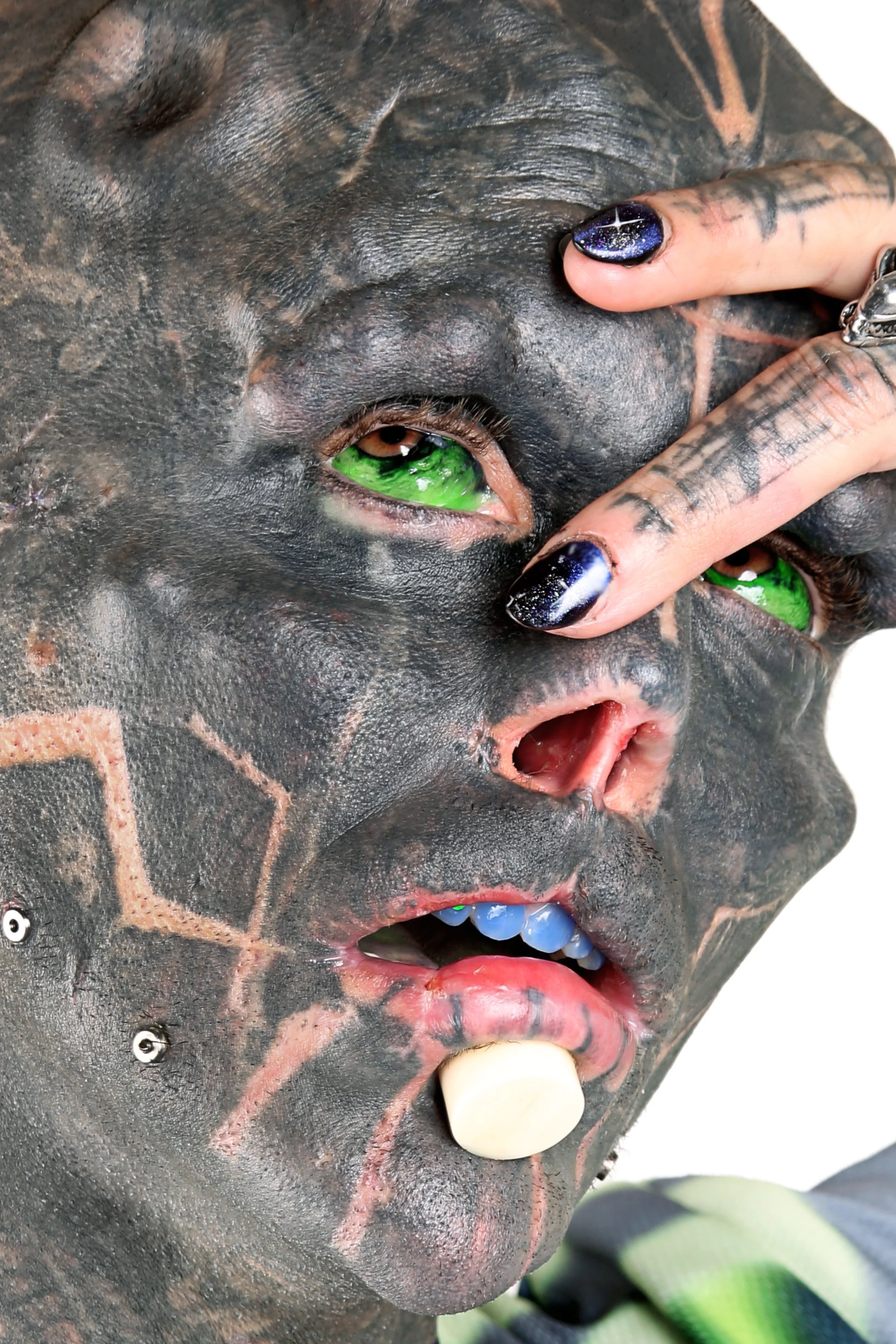 Close-up of a tattooed person's face with green eyes, multiple piercings, and their finger with dark nail polish touching their eye.