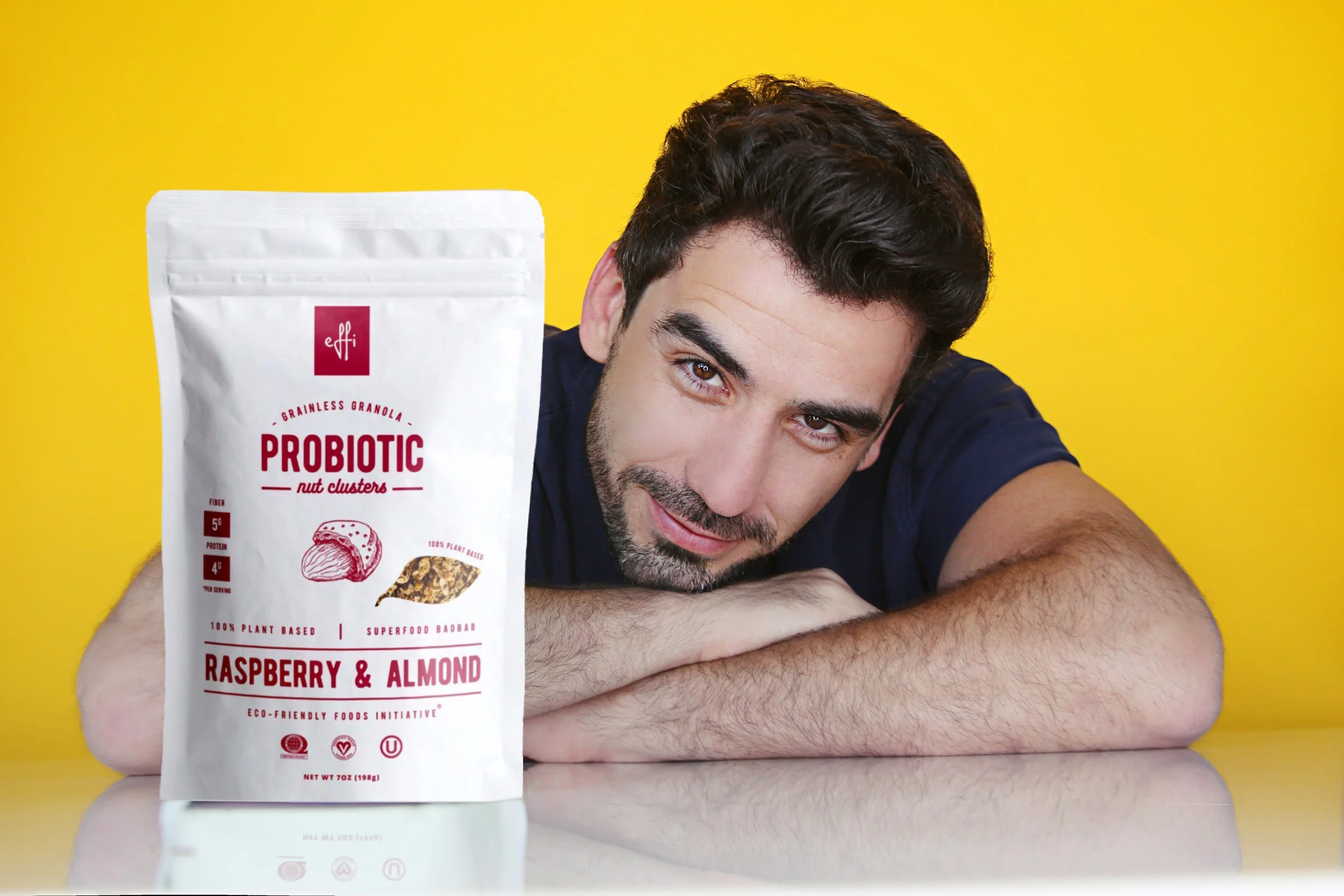 A man with dark curly hair and a beard is lying on his side on a white surface with a bright yellow background. He is smiling and resting his head and arms on his forearms. In the foreground, there is a white bag of Effi probiotic granola with raspbe