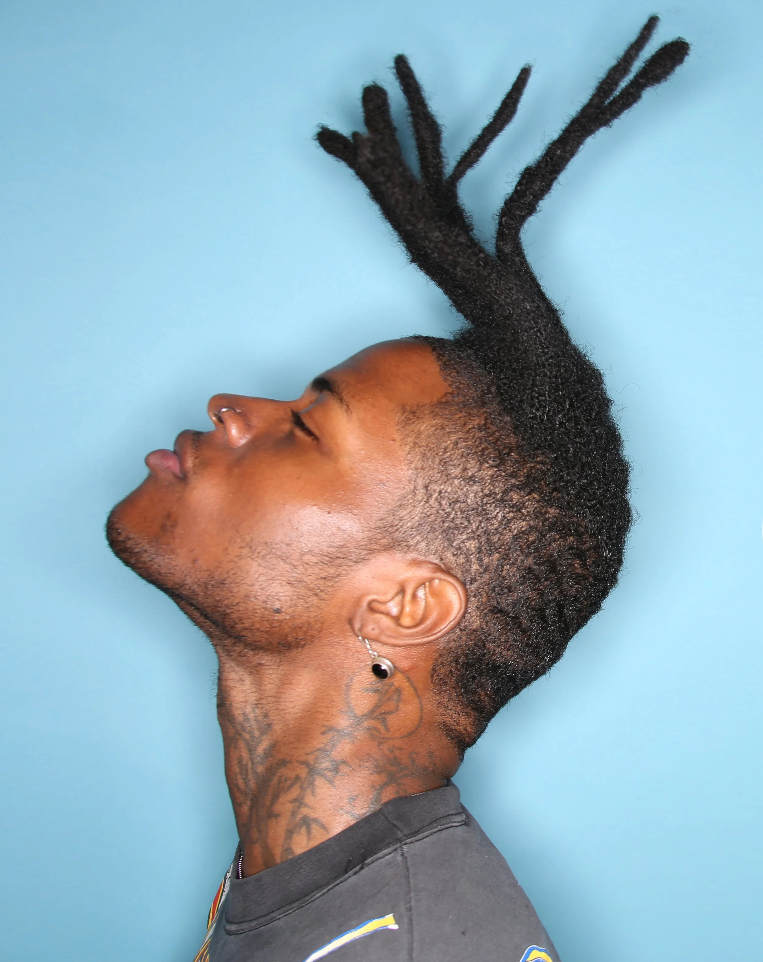 Profile of a person with dark curly hair styled in dreadlocks, wearing a black shirt with a yellow and blue emblem, and a tattoo on the neck, against a light blue background.