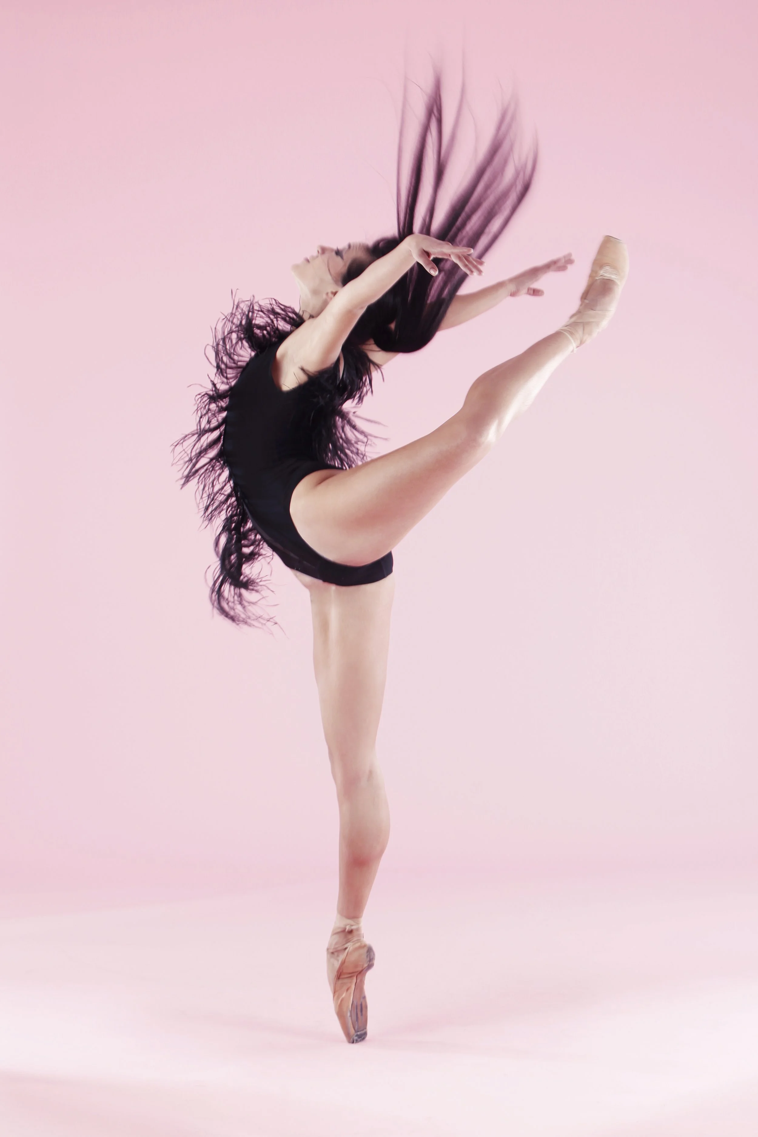 A ballet dancer in a black leotard performing a high standing split on pointe against a pink background.