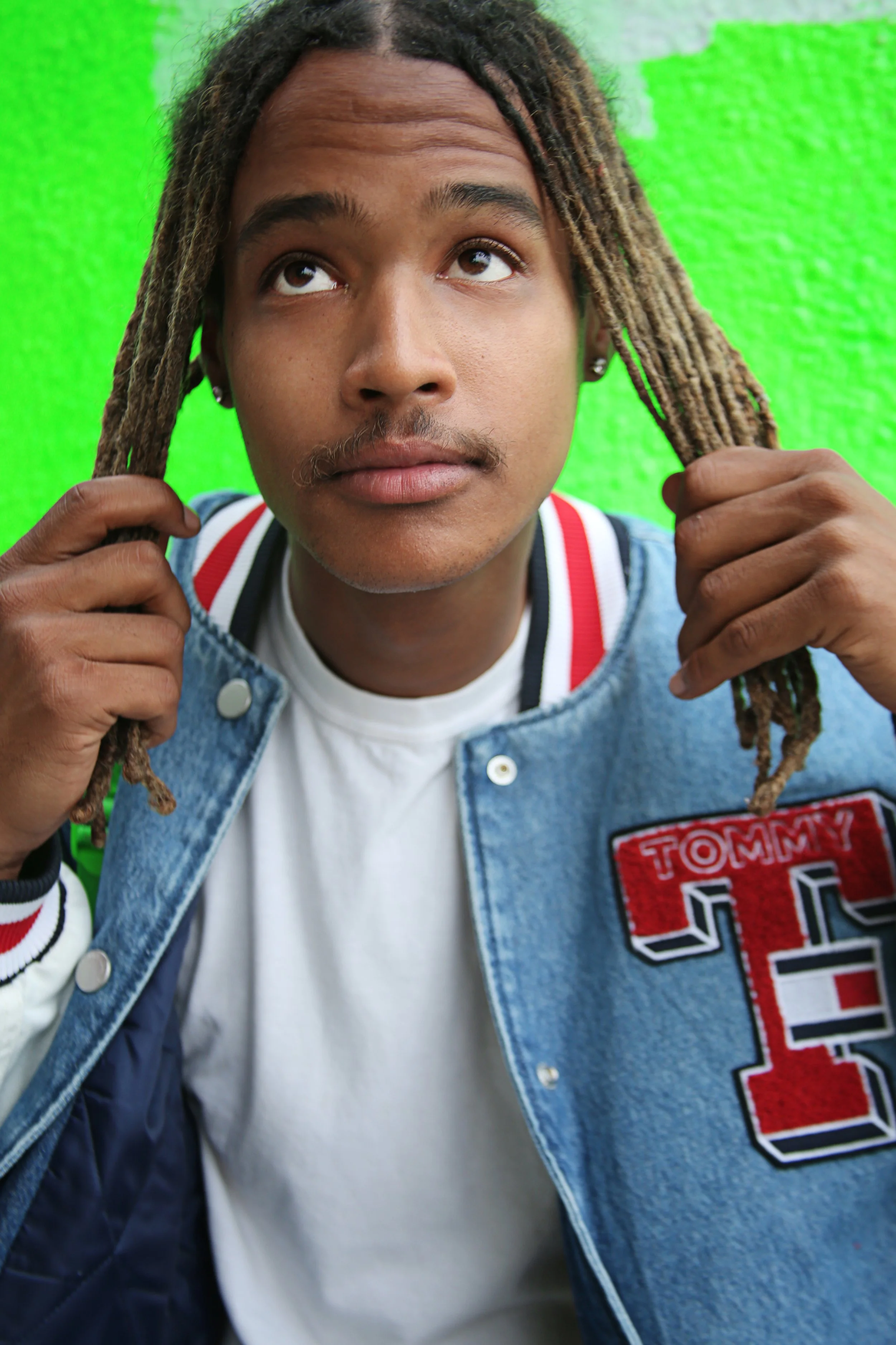Close-up of a young man with dreadlocks, wearing a denim jacket with patches, holding his hair with both hands, against a bright green background.