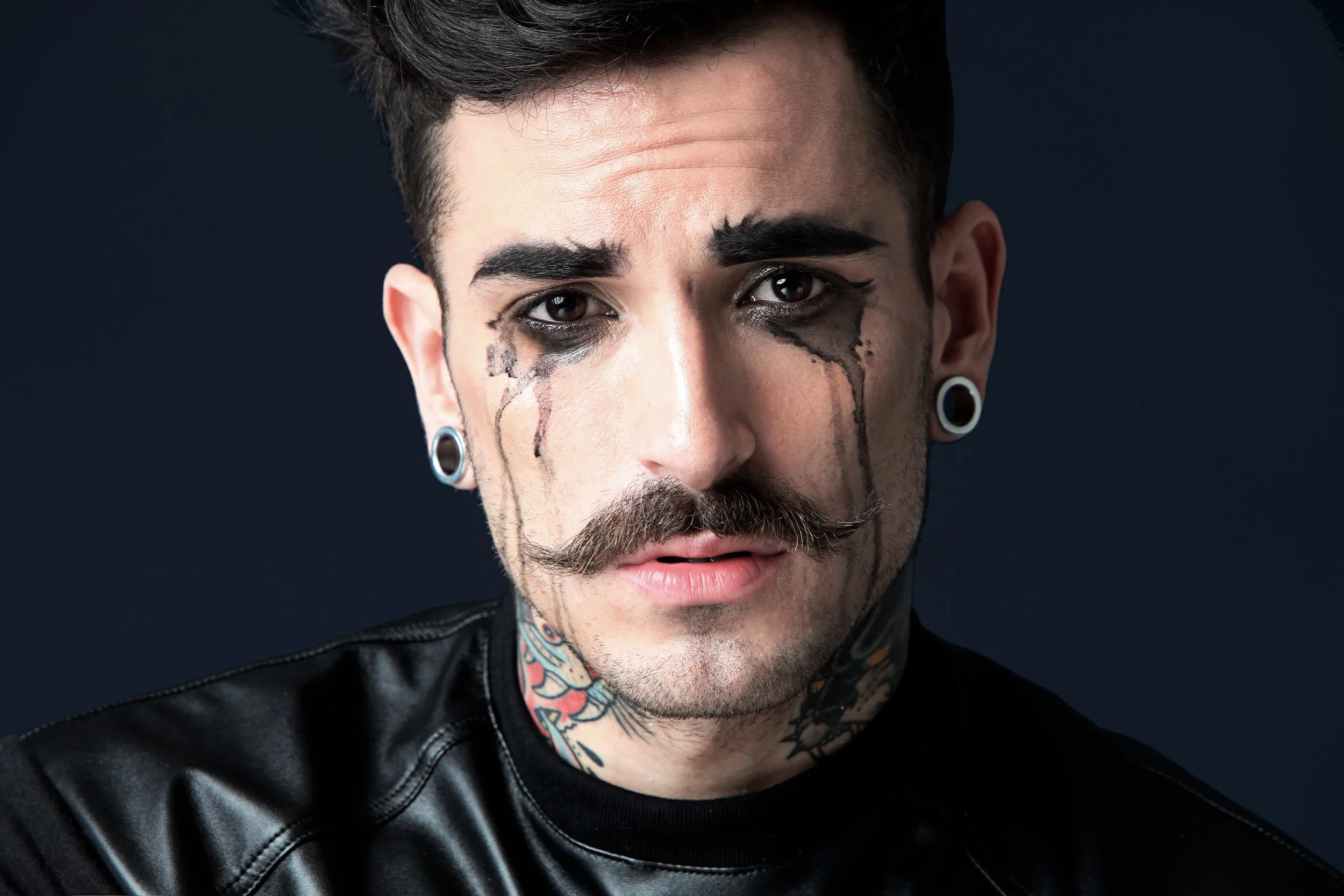 Close-up of a young man with dark hair, tattoos on his neck, wearing large plugs in his ears, and smudged black makeup around his eyes, with a serious expression against a dark background.