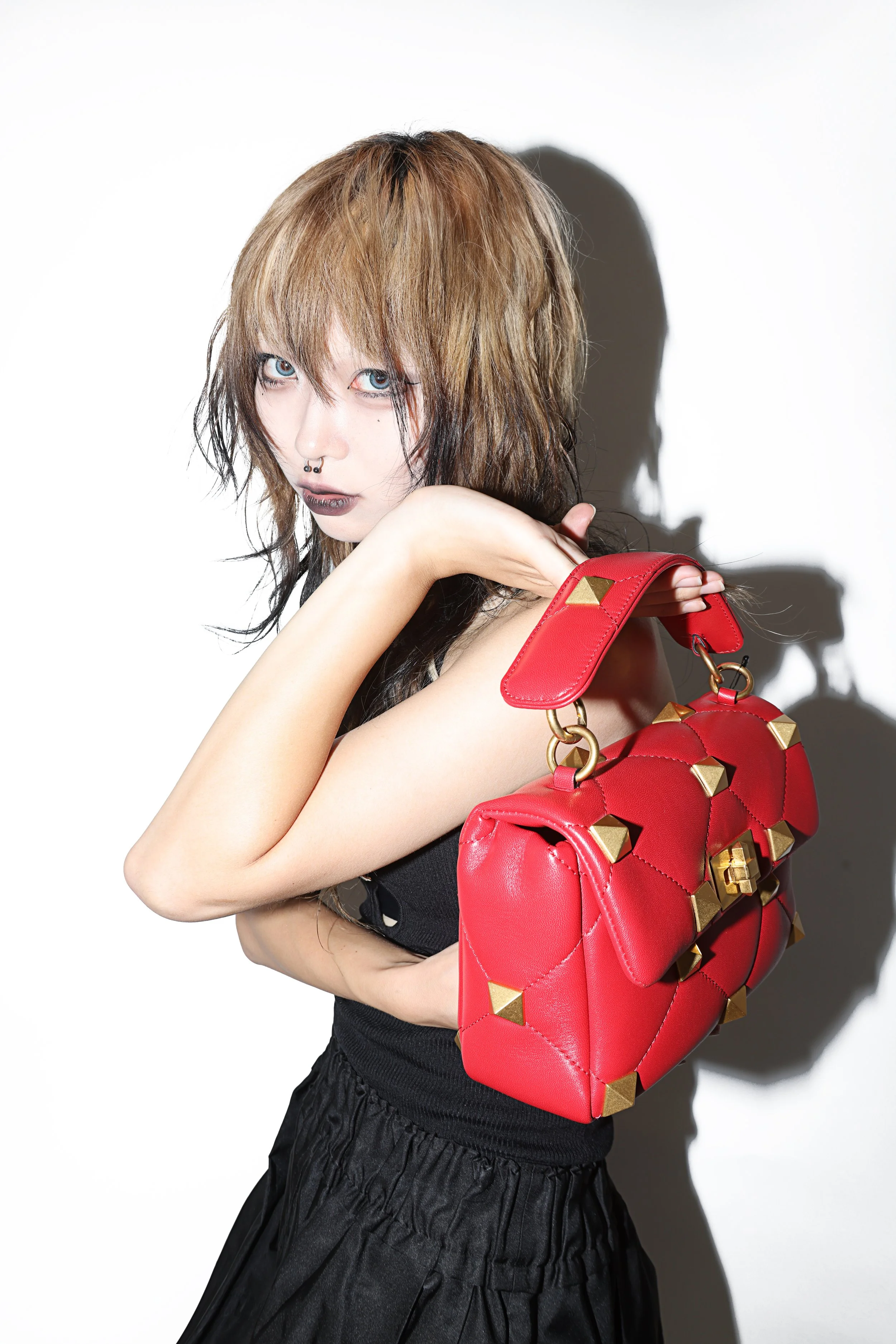 A woman with shoulder-length brown hair and blue eyes poses against a white background. She has dark lipstick and a septum piercing, and she is holding a red handbag with gold studs over her shoulder. She is wearing a black top and a black skirt.