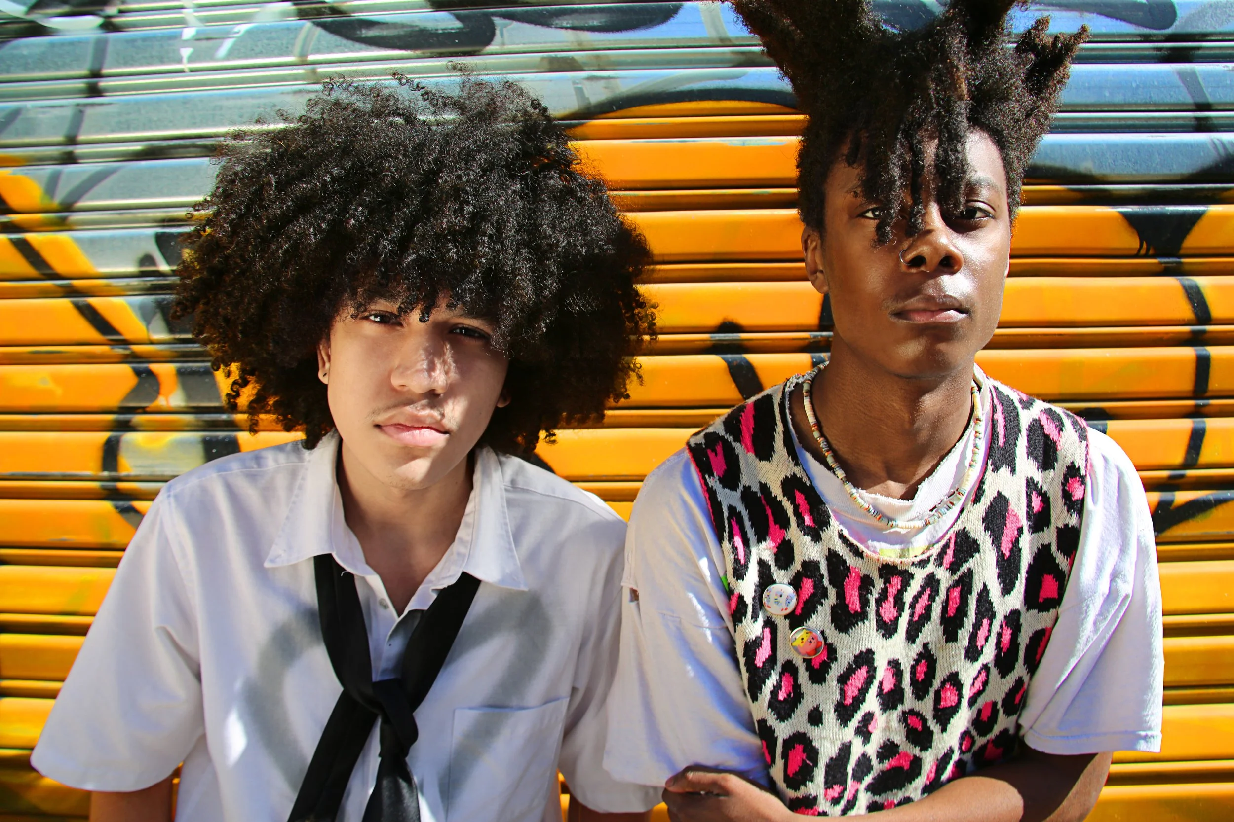 Two young people with curly hair sitting against a colorful graffiti wall, one wearing a white shirt and the other wearing a leopard print vest with buttons and pins.
