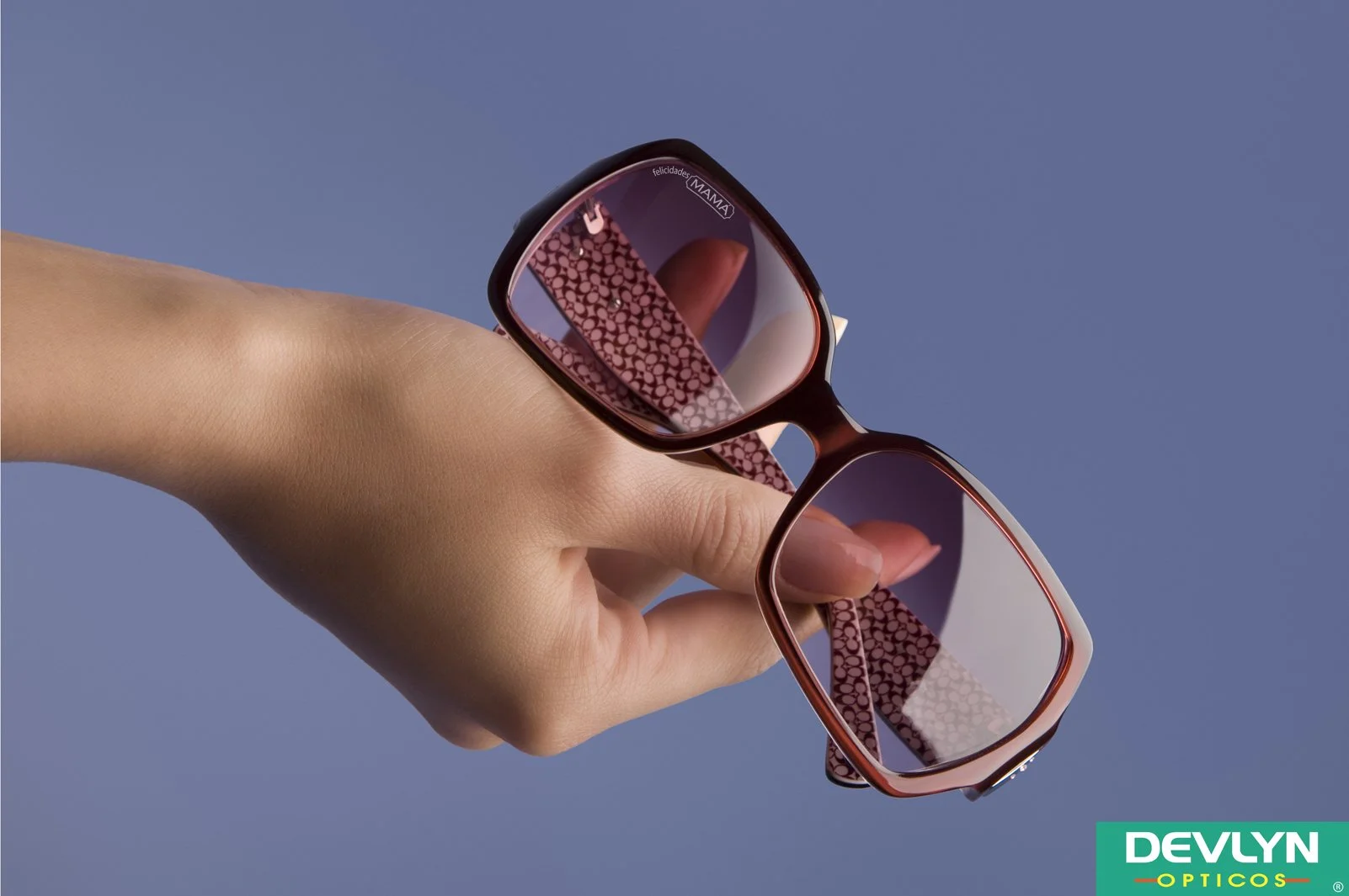 A hand holding a pair of sunglasses with pink patterned temples against a blue background. The lenses have reflections and a small sticker with the words 'felicidades MAMA' and a logo in the top right corner.