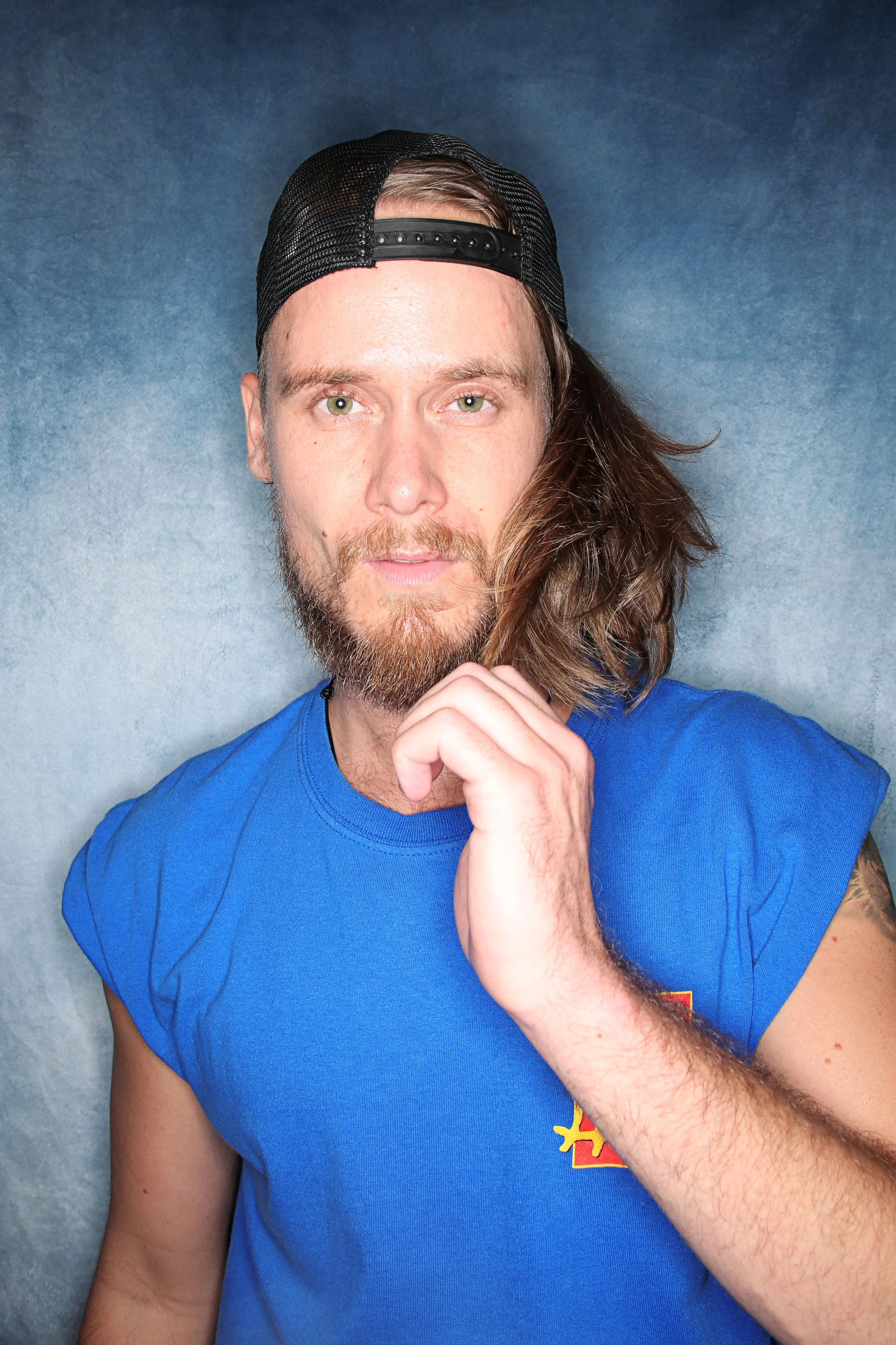 A young man with long hair, a beard, and blue eyes wearing a backwards black baseball cap and a blue T-shirt, resting his chin on his finger in front of a plain gray background.
