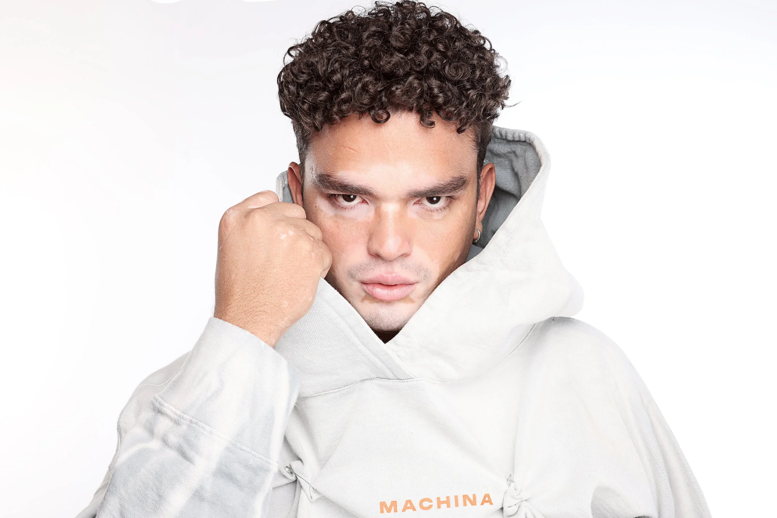 A man with curly hair wearing a white hoodie with the word 'MACHINA' printed on it, looking directly at the camera, with his right hand touching his face.