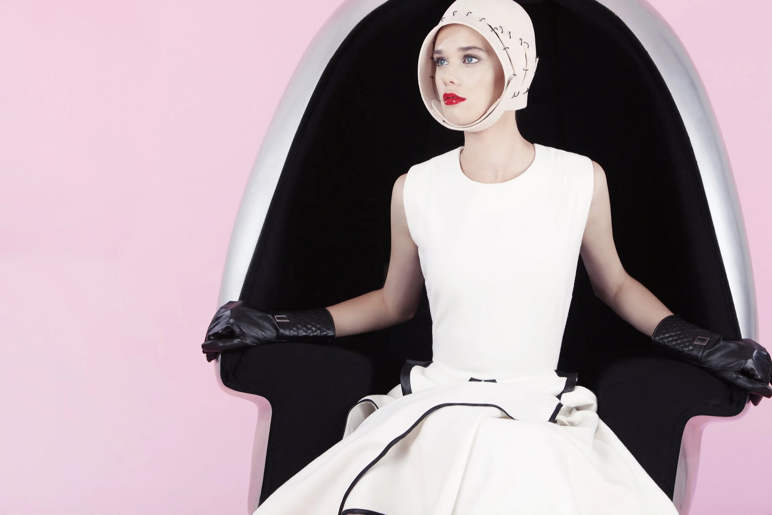 A woman with blue eyes and red lipstick wearing a white dress and a head covering, sitting inside a black pod chair with a pink background.