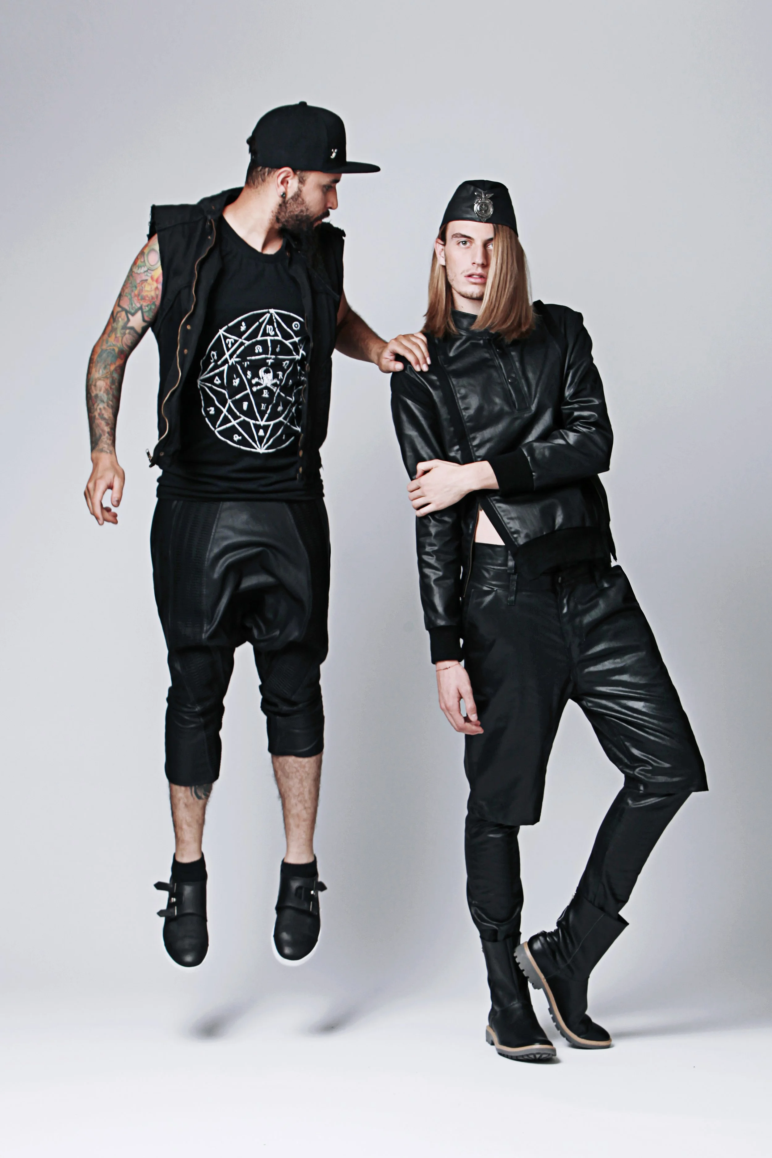 Two young men, one with tattoos and a beard wearing a black cap and sleeveless hoodie, the other with long hair wearing a black leather jacket, black pants, and boots, posing against a plain white background.