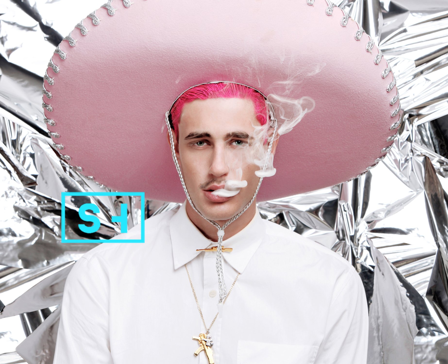 A young man with pink hair wearing a large pink sombrero, smoking, dressed in a white shirt with multiple necklaces, standing in front of a shiny metallic background.