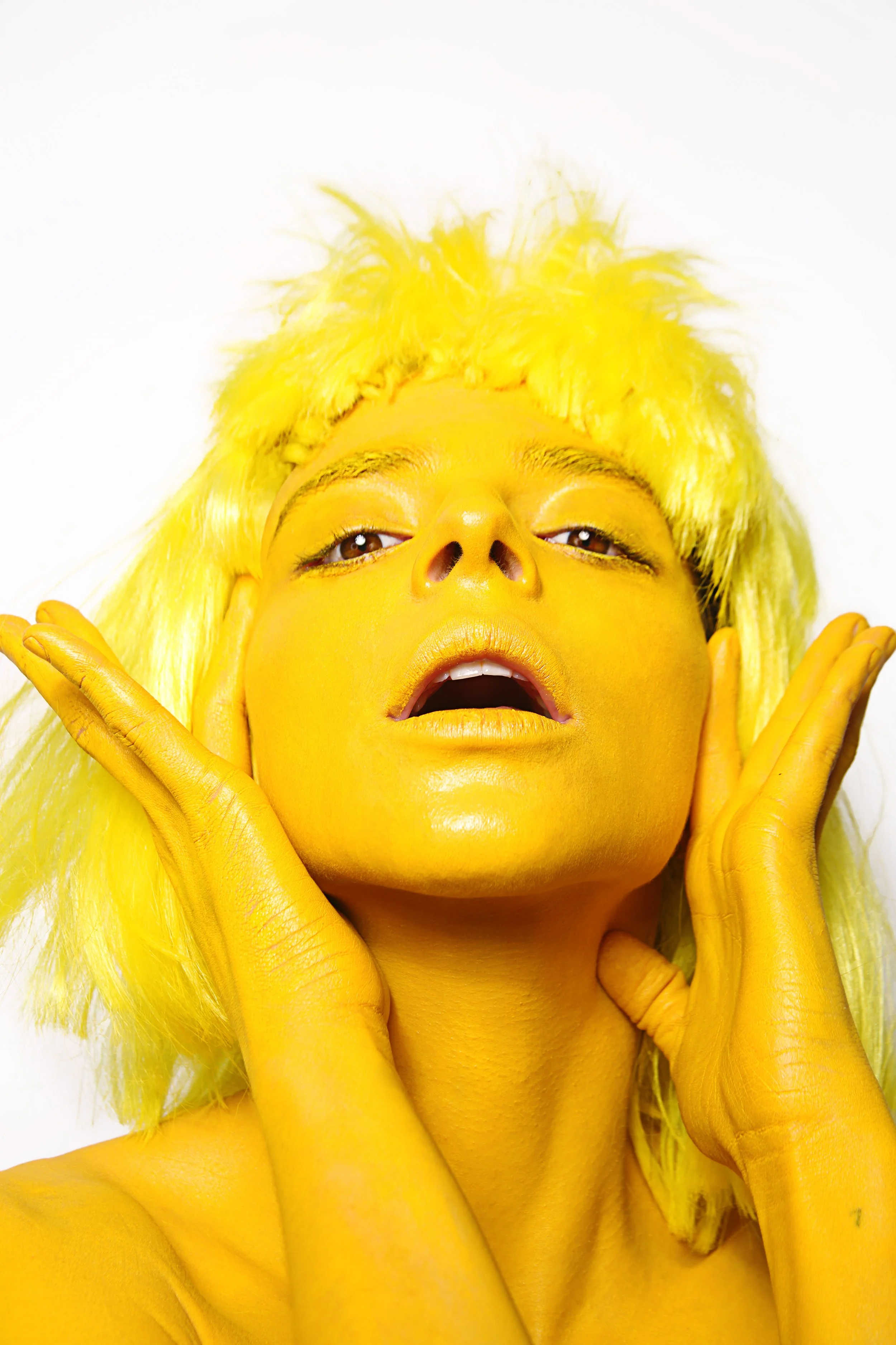 Person with bright yellow skin, yellow hair, wearing yellow gloves, and yellow makeup, posing with hands around their face against a white background.