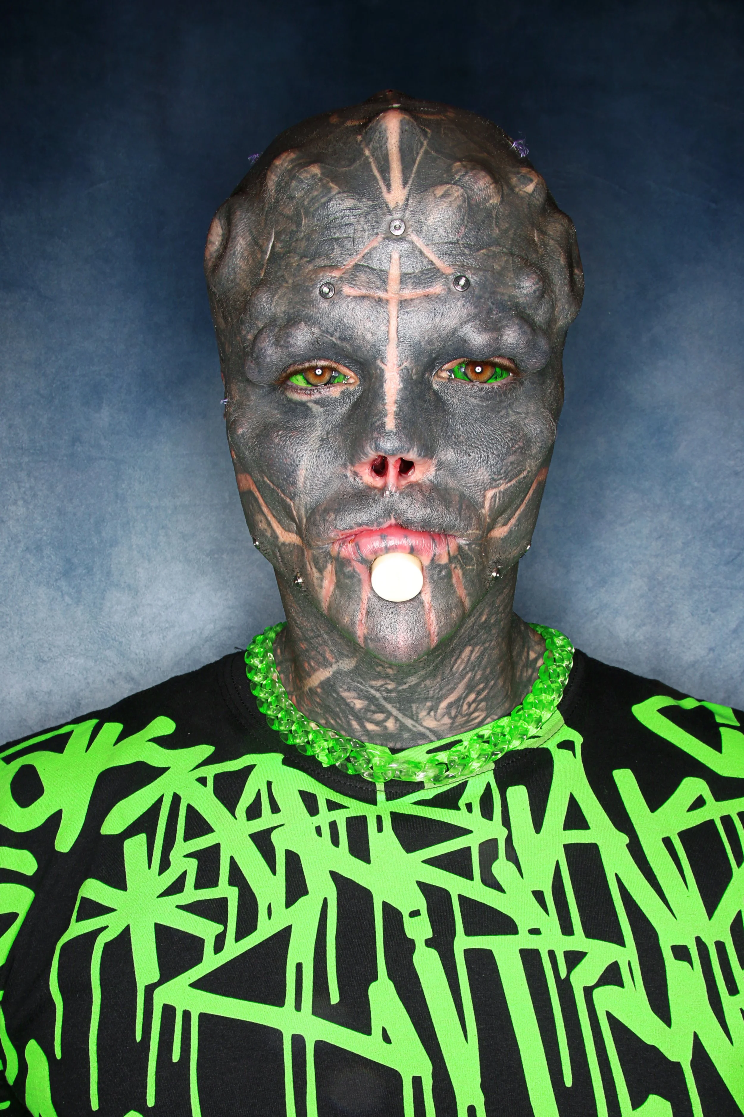 Person with face and head tattoos, wearing a black and neon green graphic shirt, a green beaded necklace, and holding a white lollipop in mouth, against a neutral background.