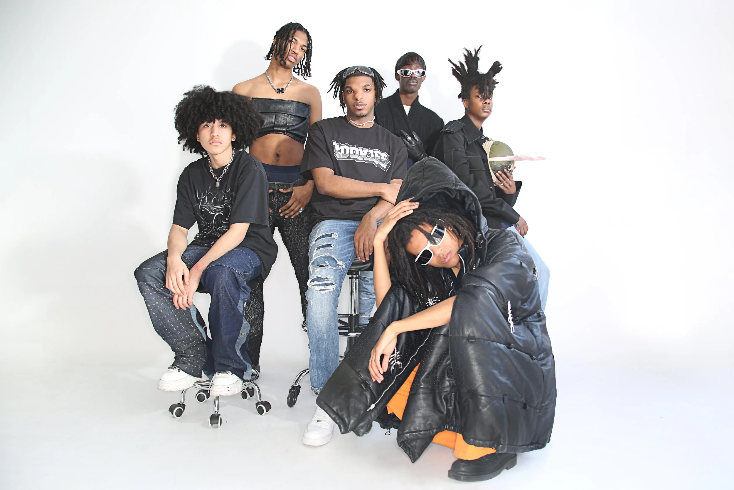 Group of six diverse young adults posing against a white background, dressed in casual and edgy fashion, with some wearing sunglasses and accessories.