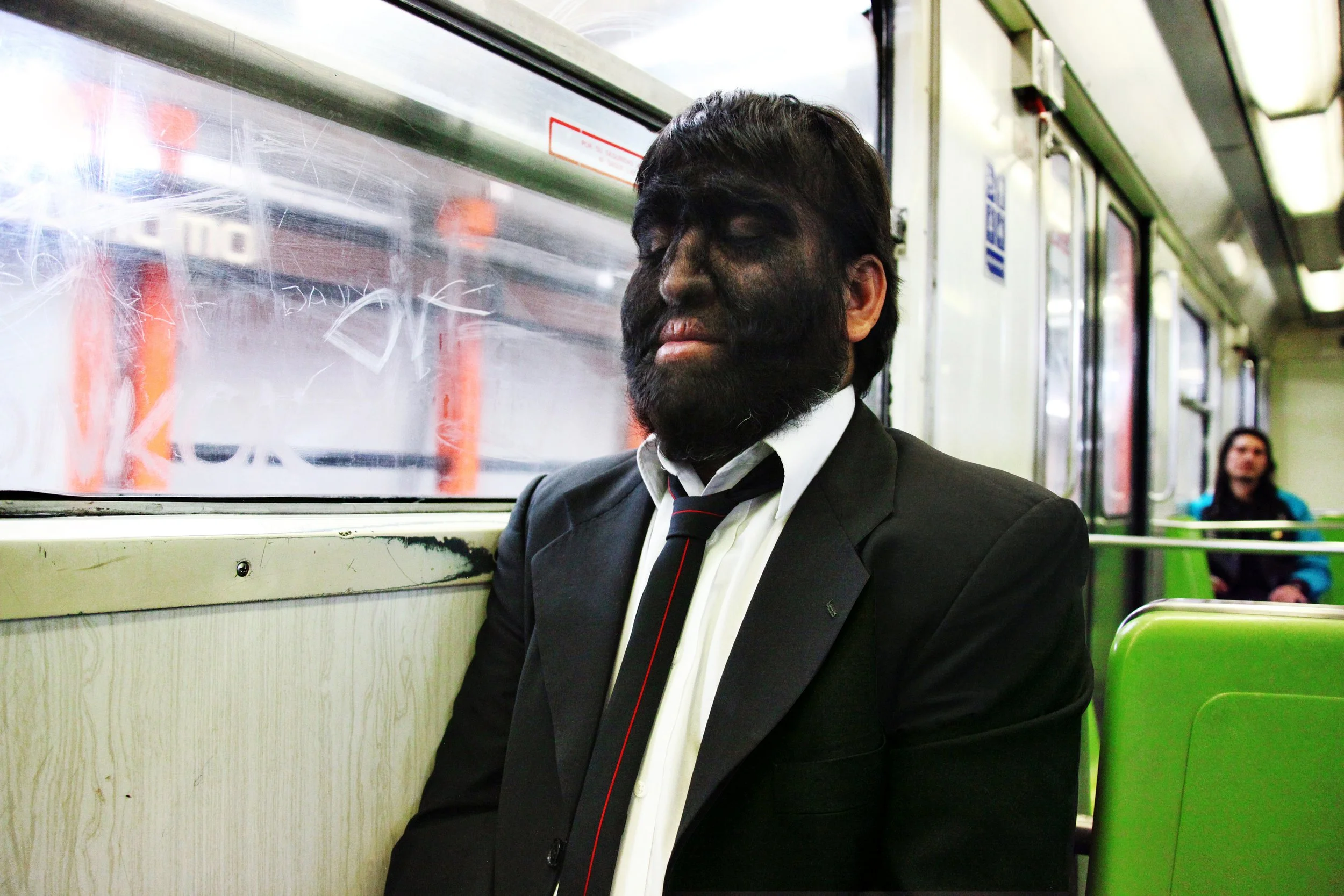 A man with closed eyes and a full beard, dressed in a black suit with a white shirt and black tie with red stripes, is sitting on a green seat on a bus or train. He appears to be resting or sleeping.