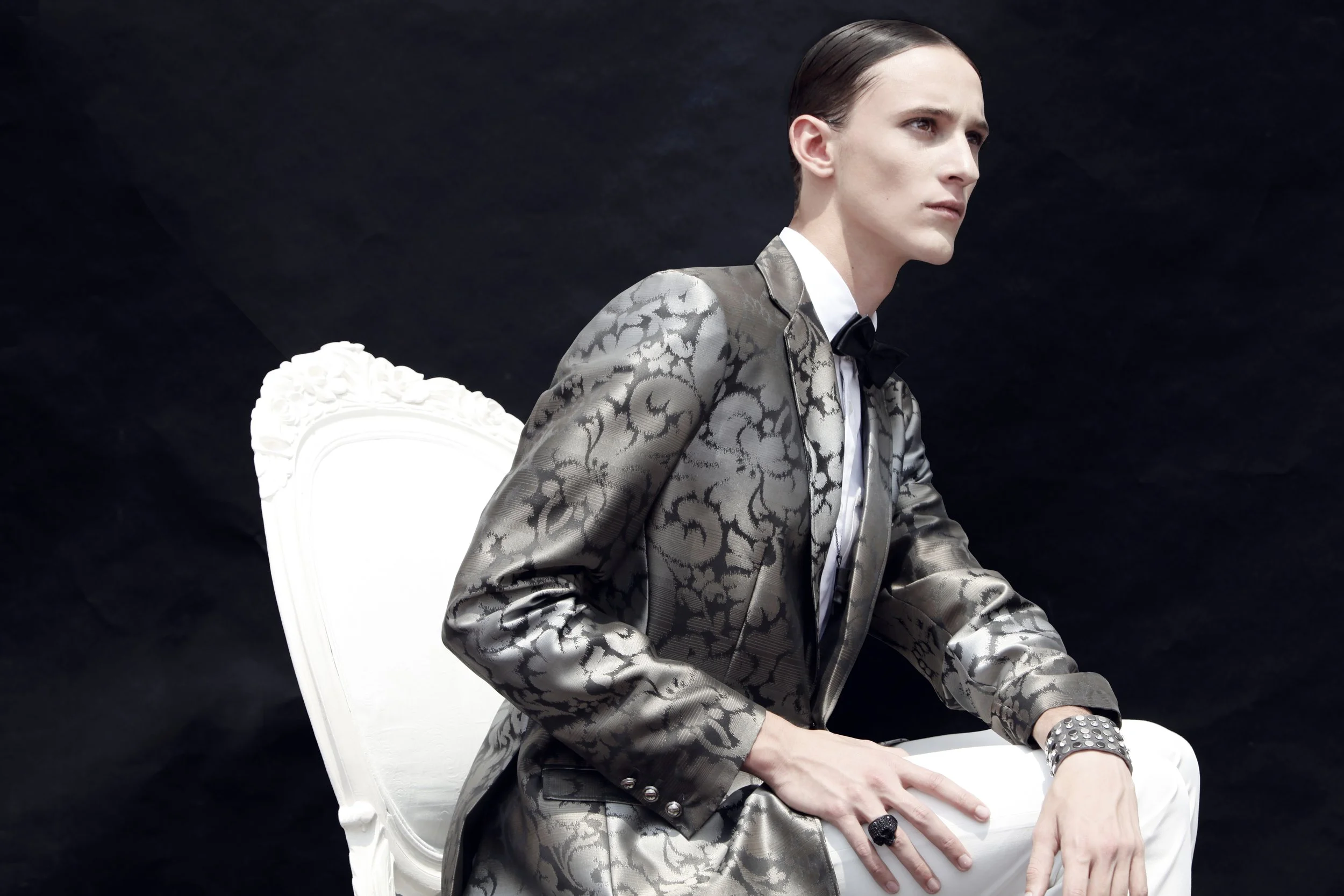 A man with slicked-back hair, wearing a shiny patterned suit with a black bow tie, sitting on a white ornate chair against a black background.