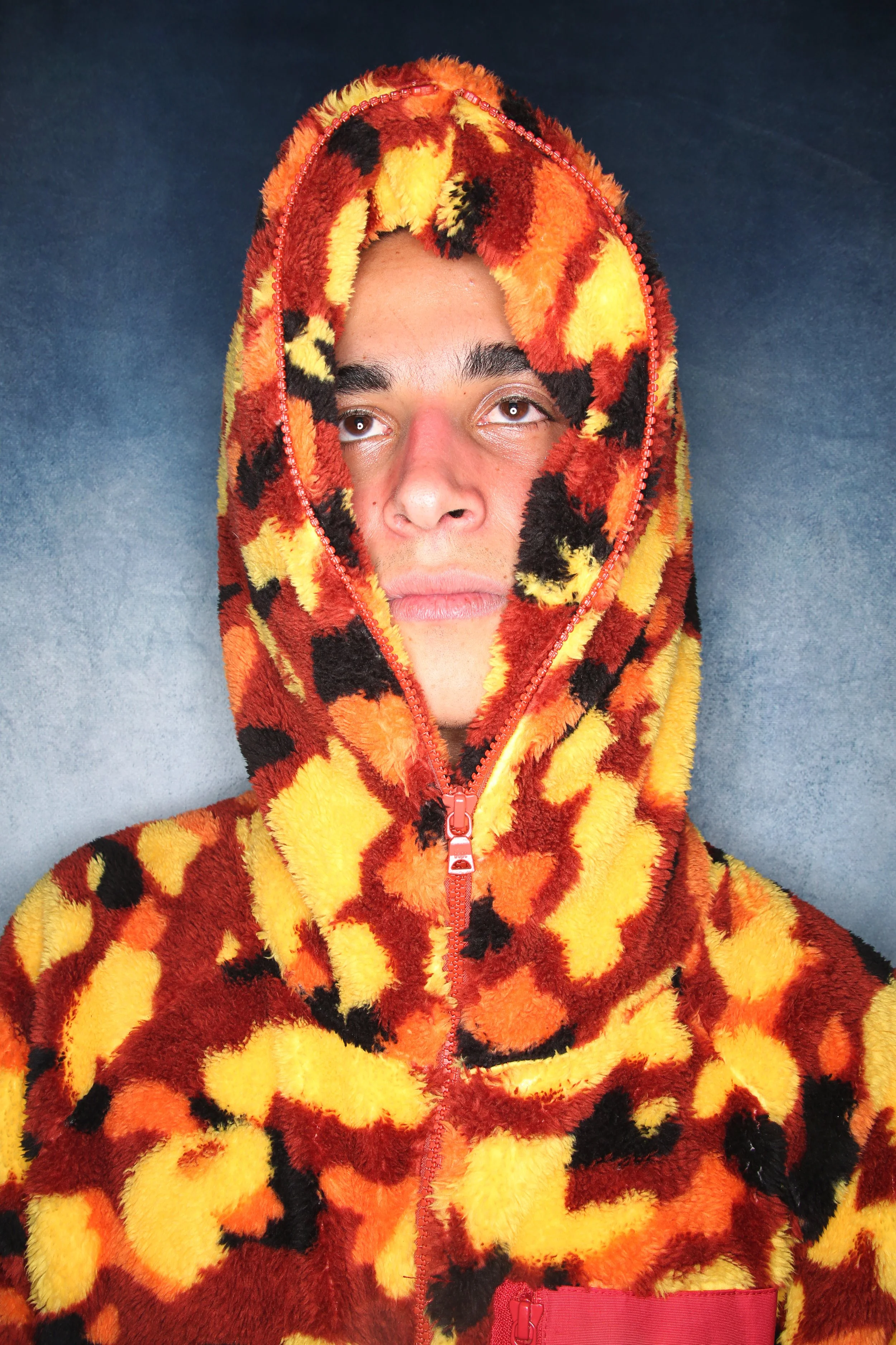A man lying on a blue surface wearing a fuzzy, tiger-print hoodie with a zipper, featuring black, orange, and yellow patterns.