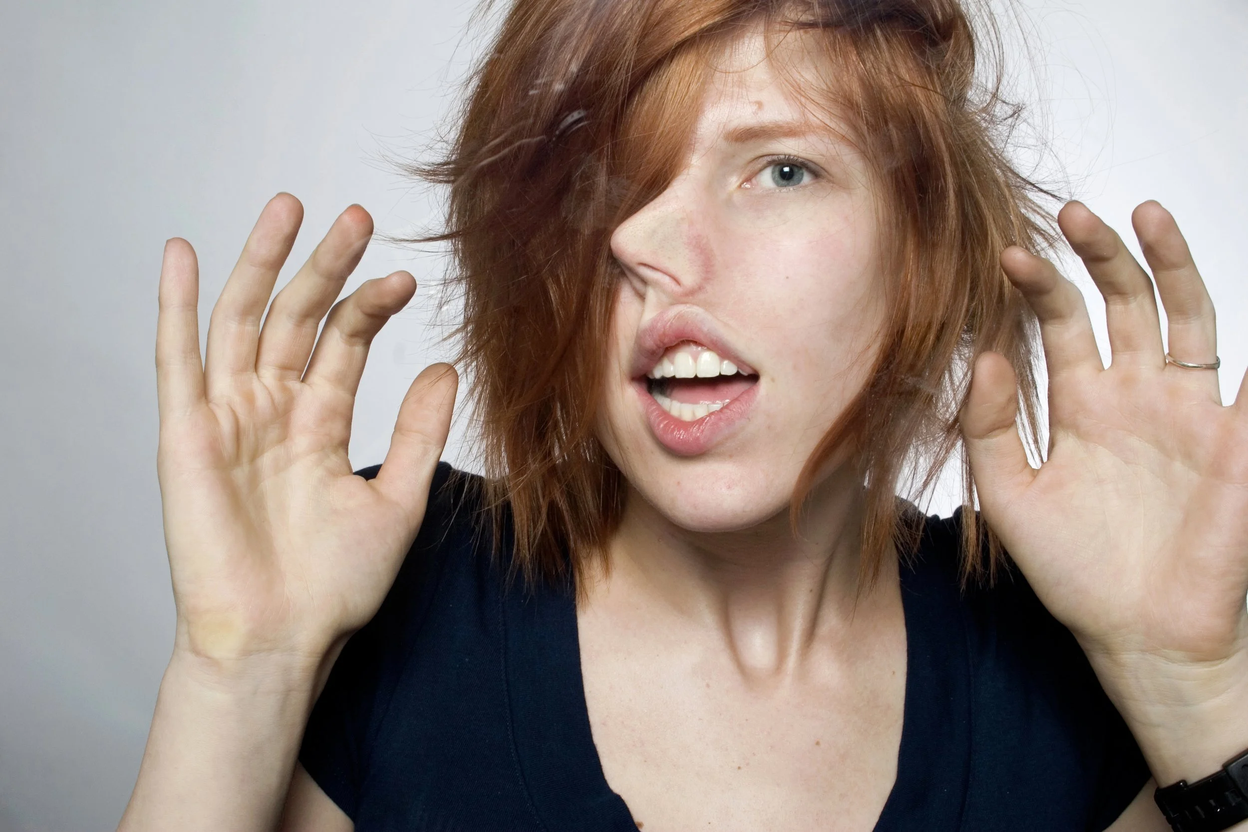 A young woman with red hair is making a surprised facial expression with her mouth open and eyes wide. She is holding her hands up near her face with fingers spread apart.