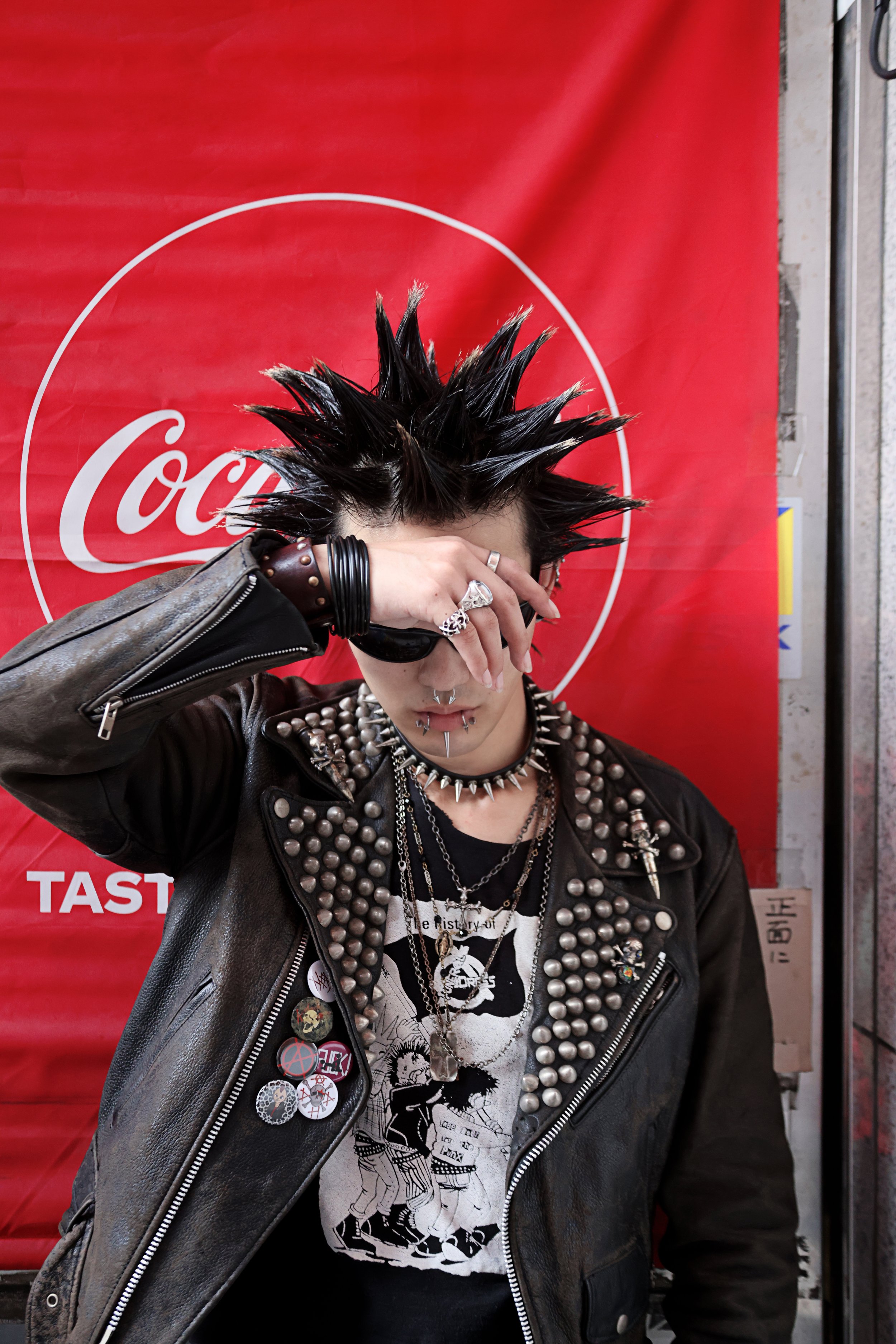 Person with spiky black hair wearing a leather jacket with metal studs, lots of pins and buttons, jewelry including rings, necklaces, and earrings, covering their face with their hand in front of a red Coca-Cola backdrop.