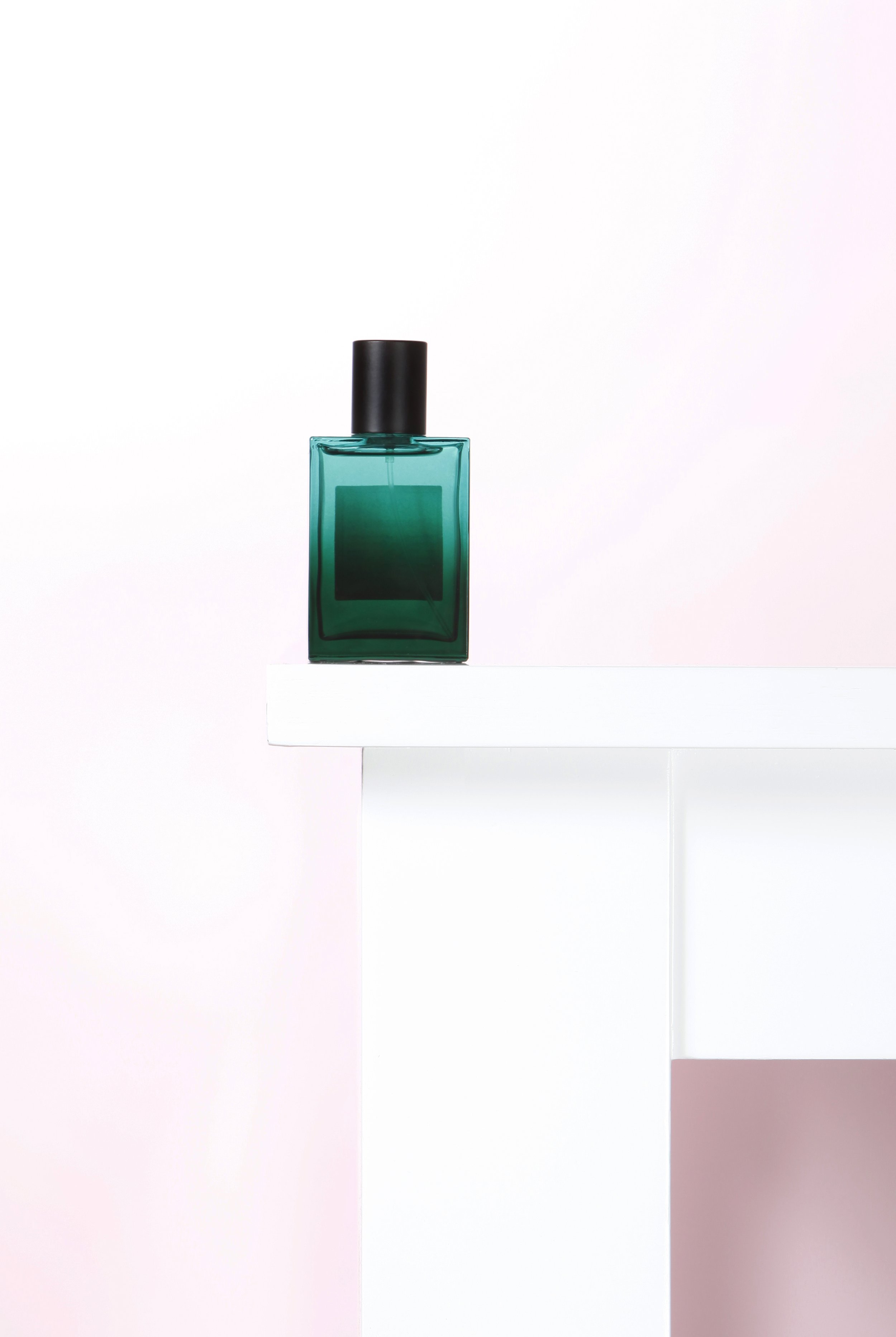 A teal perfume bottle with a black cap is placed on a white platform against a white and soft pink gradient background.