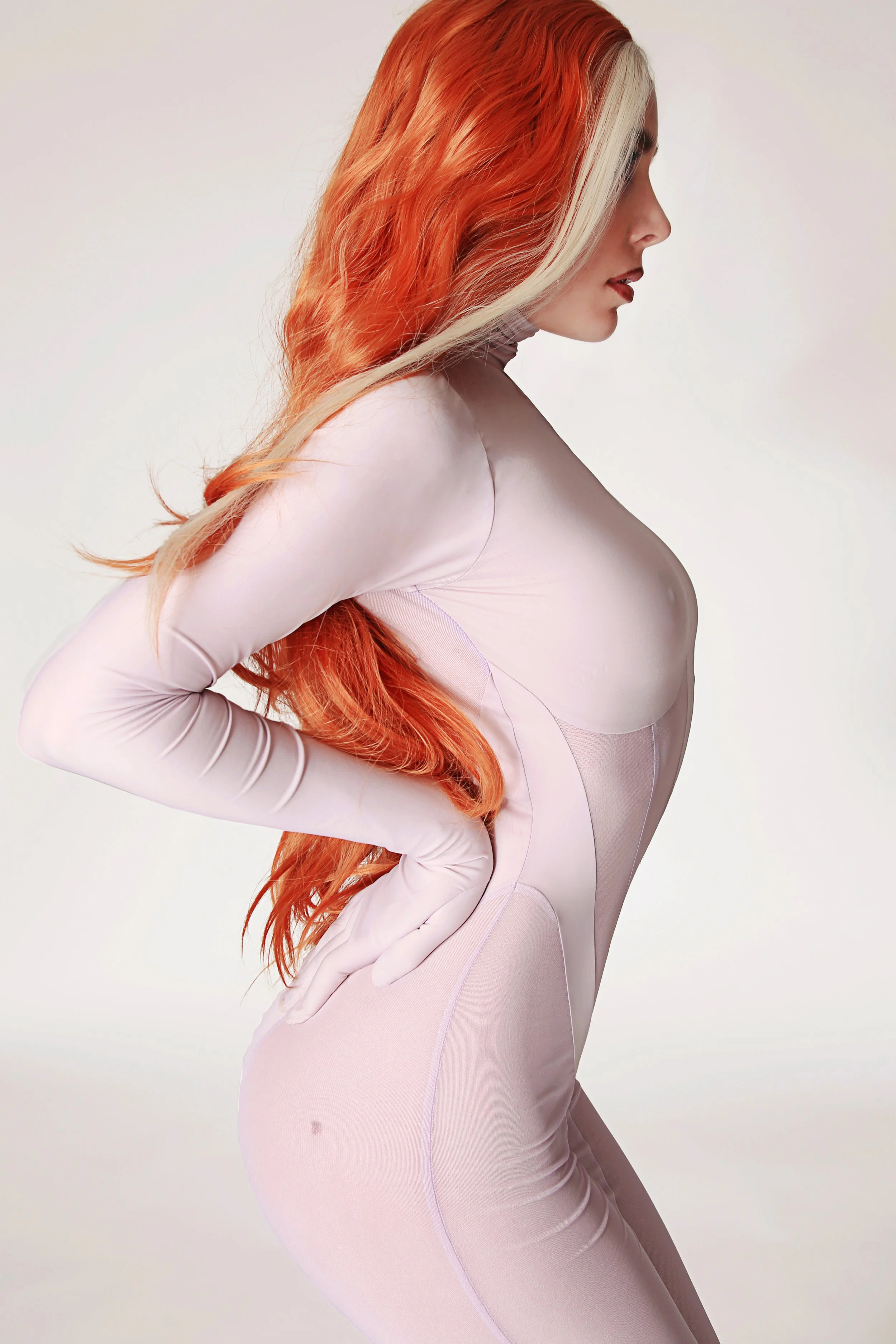Side profile of a woman with long, wavy, ombre hair transitioning from blonde to vibrant red, wearing a pale pink, fitted, long-sleeve bodysuit, standing against a plain white background.