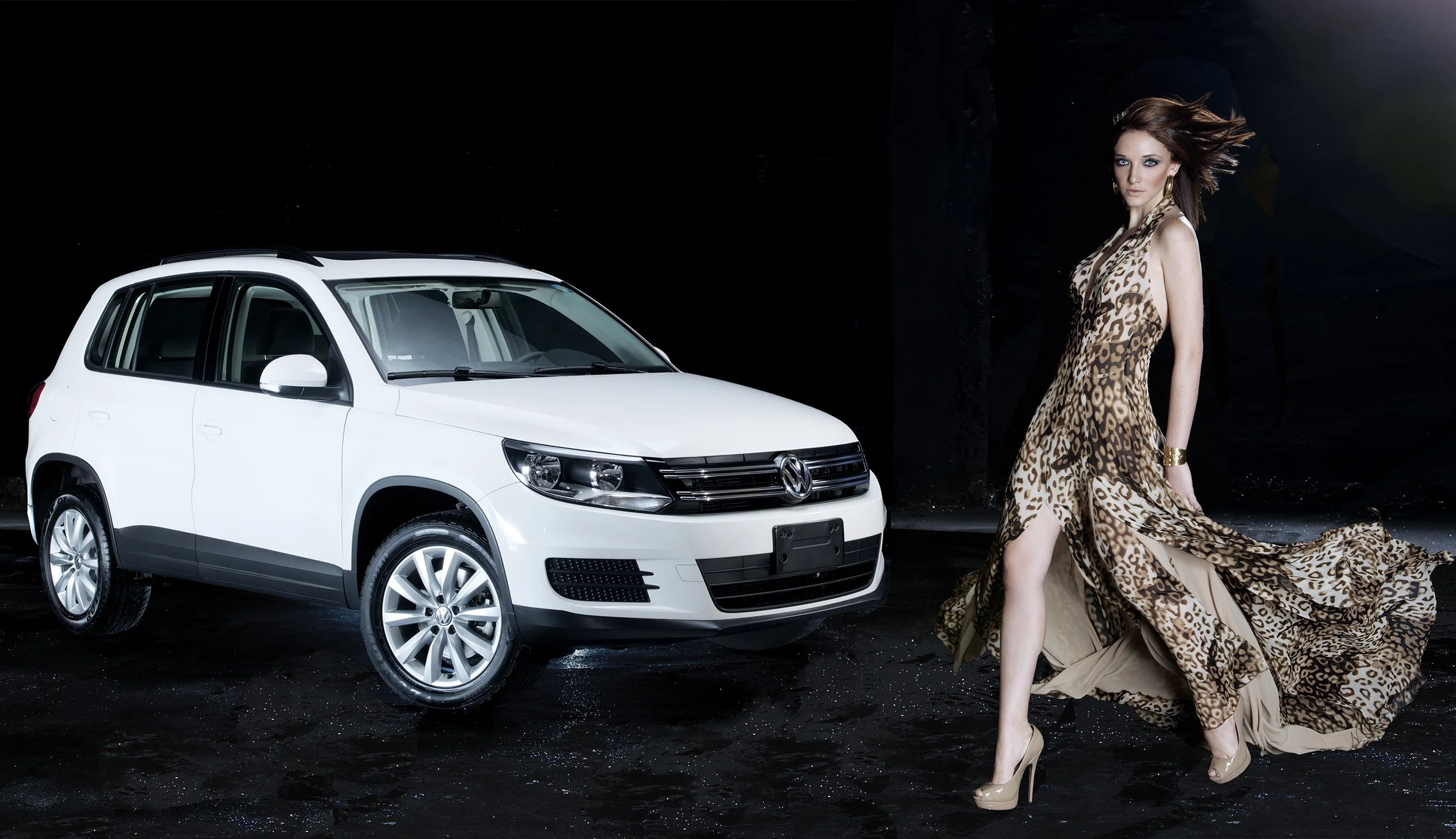 A woman in a leopard print dress with high heels and gold jewelry walking in front of a white Volkswagen car at night, with dark surroundings.
