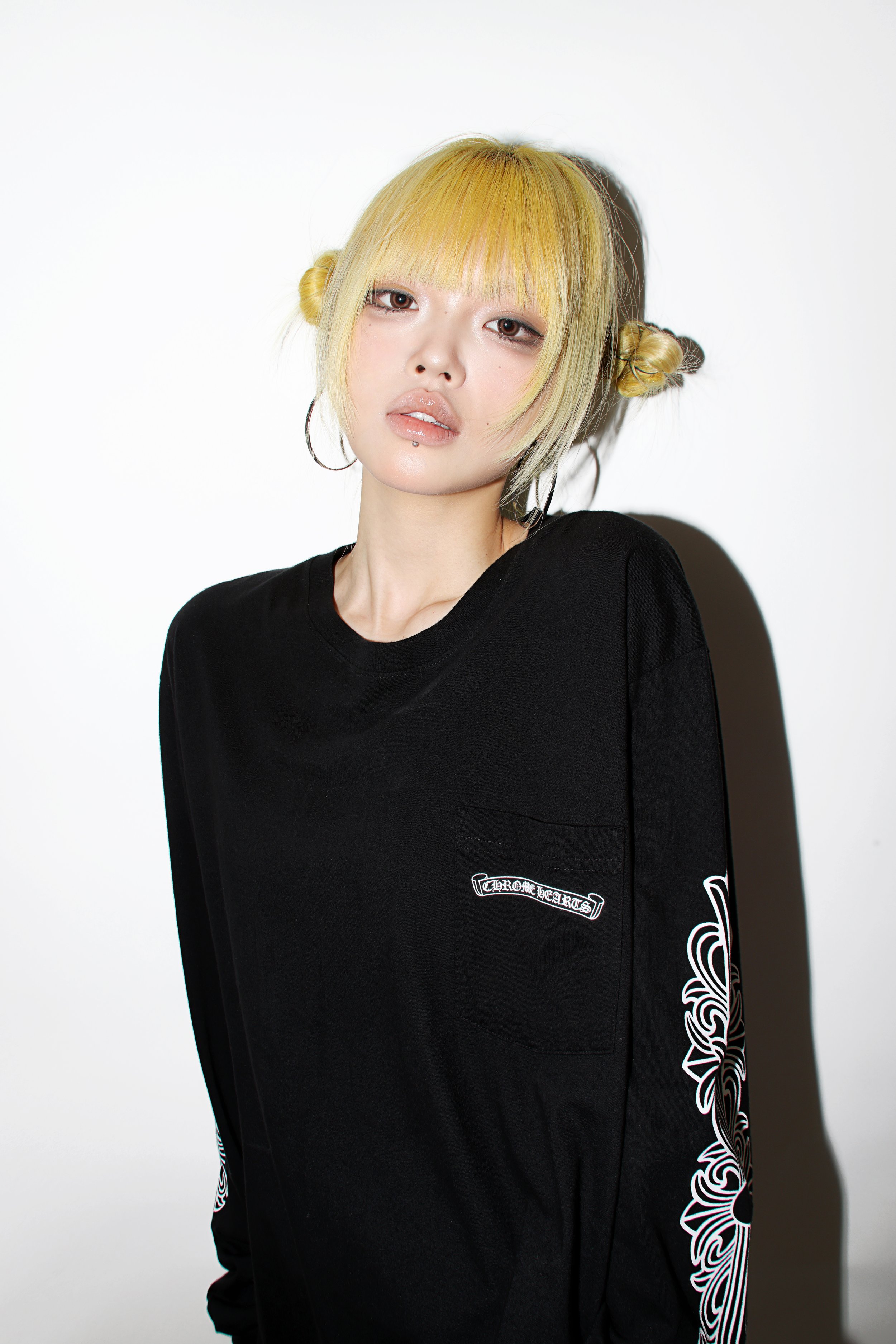 Person with yellow hair styled in buns wearing a black long-sleeve shirt with white graphic embroidery on the sleeves, standing against a white background.