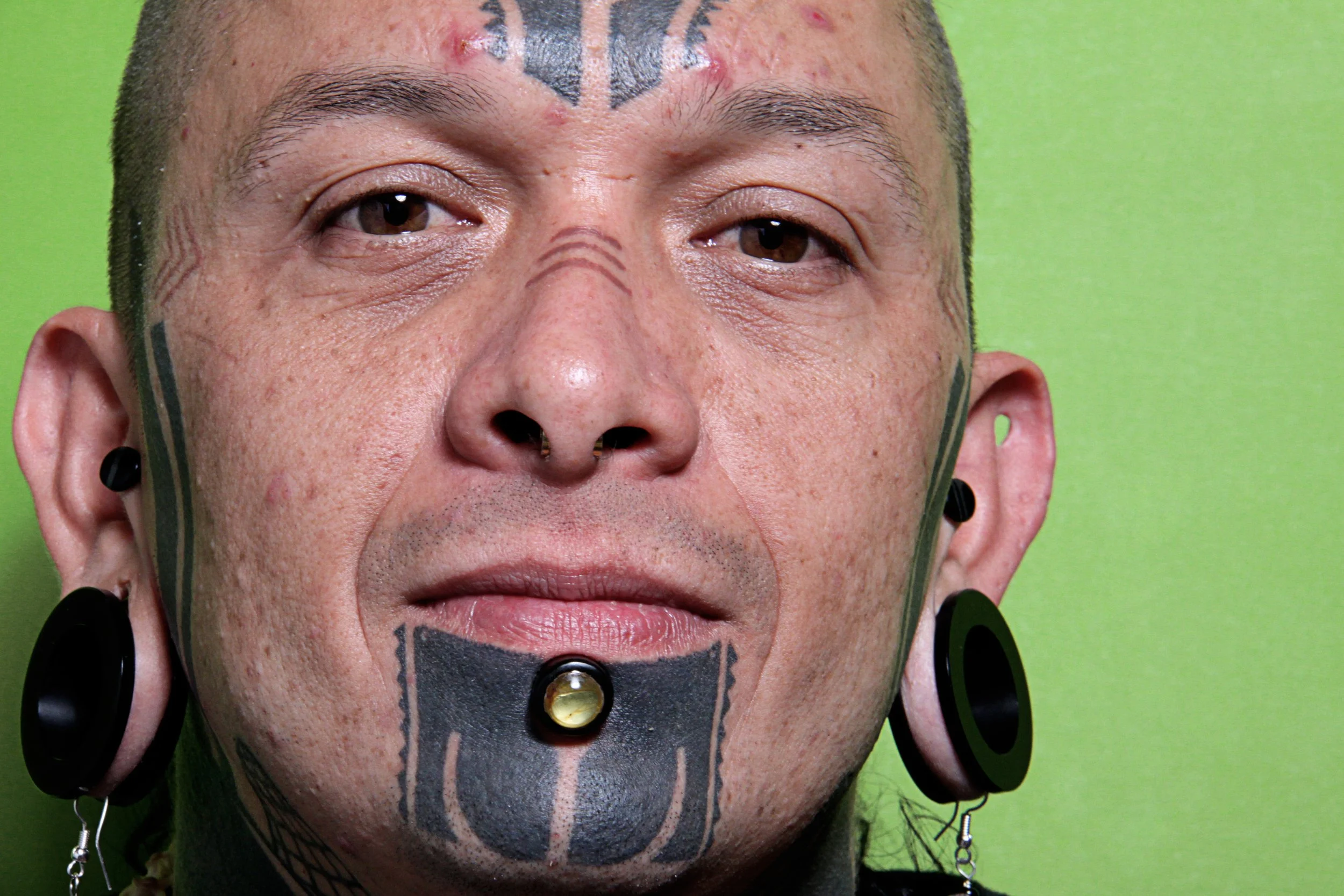 Close-up of a person's face with tattoos, piercings, and jewelry, set against a green background.
