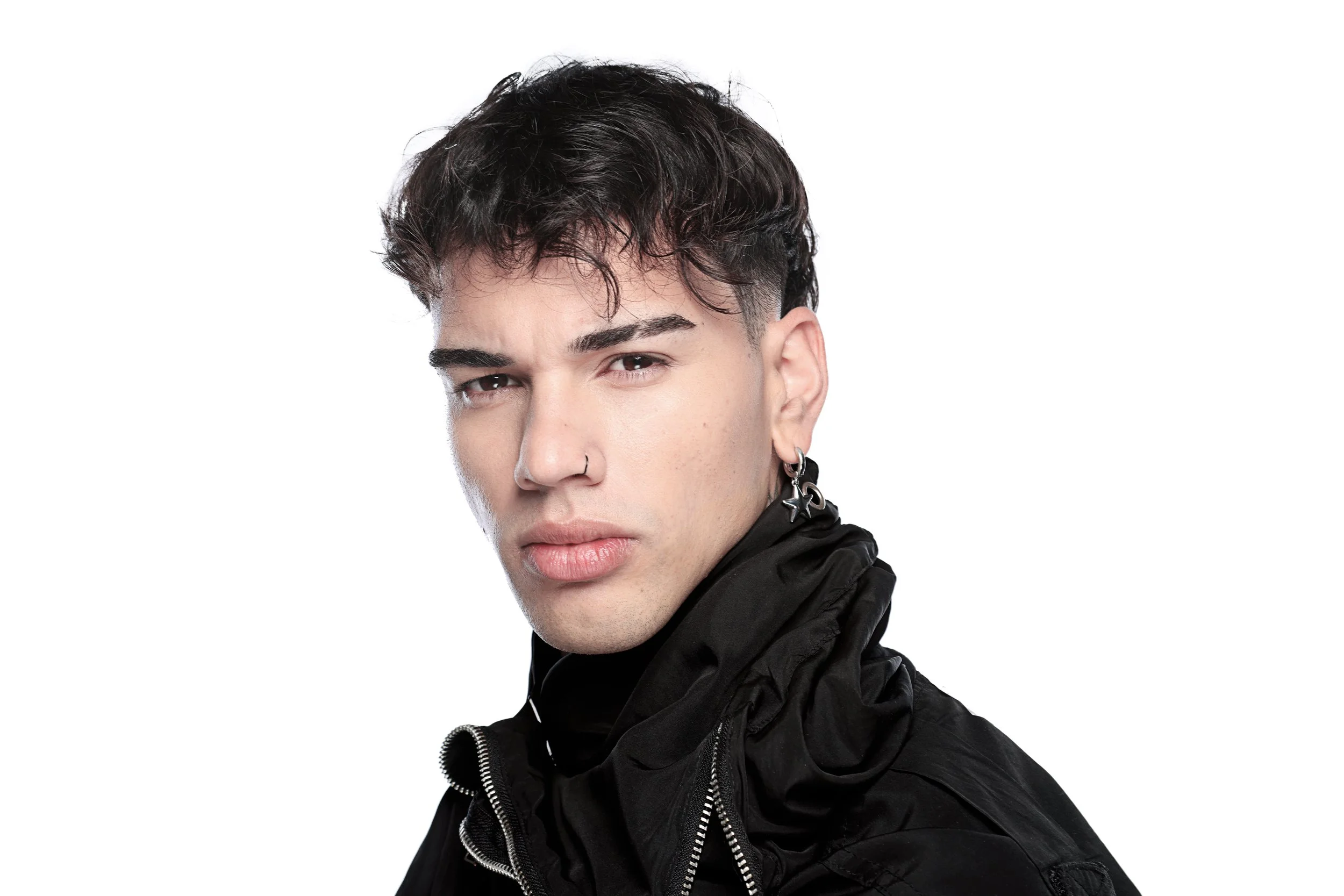 Portrait of a young man with dark, wavy hair, earrings, and a nose ring, wearing a black hoodie, against a white background.