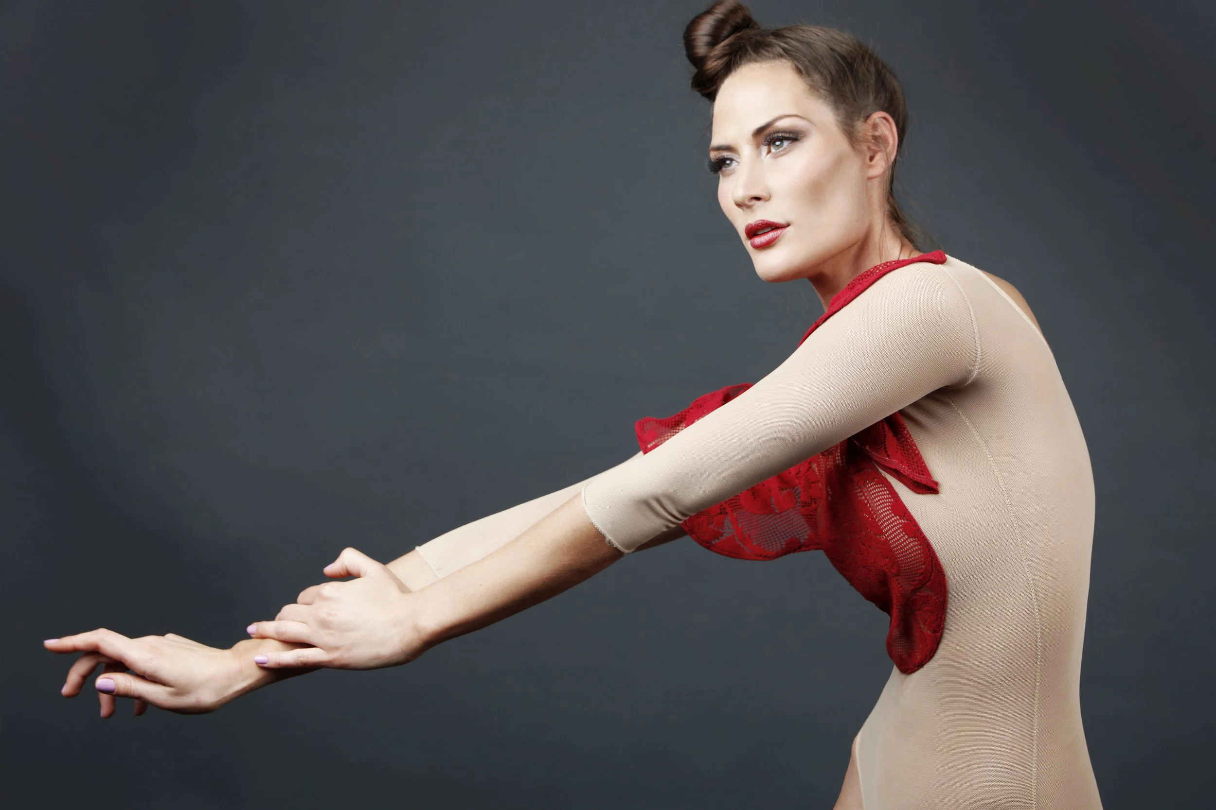 A woman with light skin, red lipstick, and styled brown hair with a bun, wearing a beige top with red lace accents, is stretching her arms forward against a dark background.