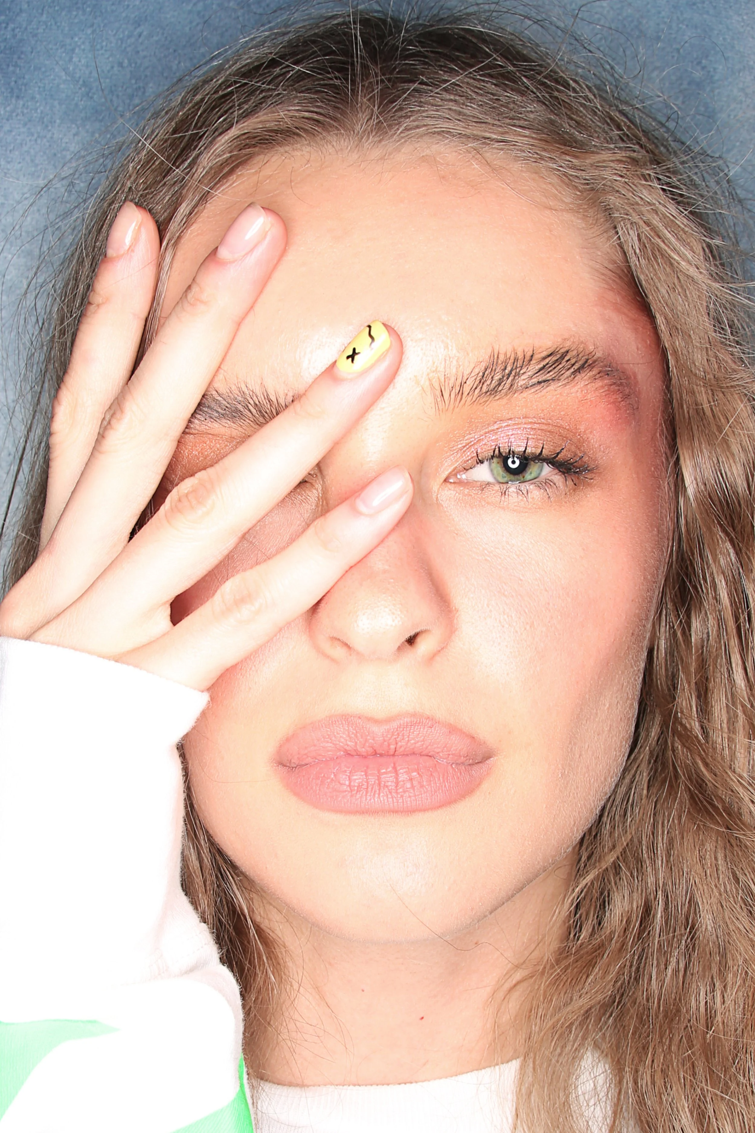 Close-up of a young woman with wavy brown hair and green eyes, partially covering her face with her hand that has a yellow nail with a black smiley face design.