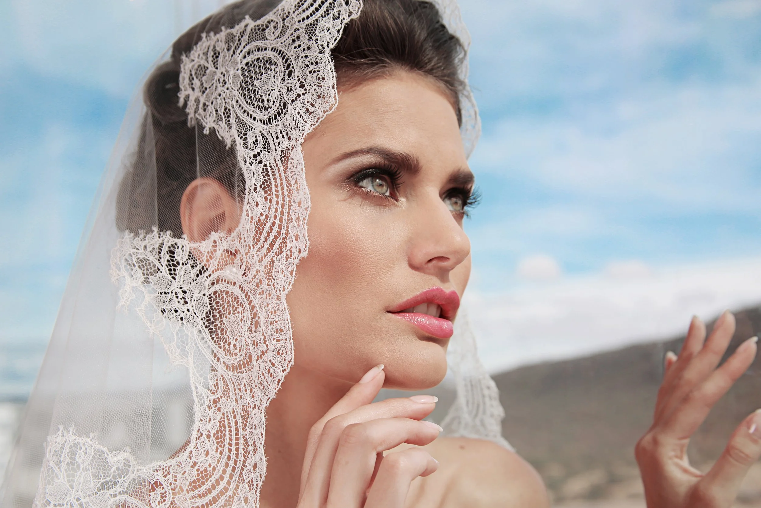 A woman with makeup and a lace veil on her head, looking at her reflection, possibly outside at a beach or scenic area.
