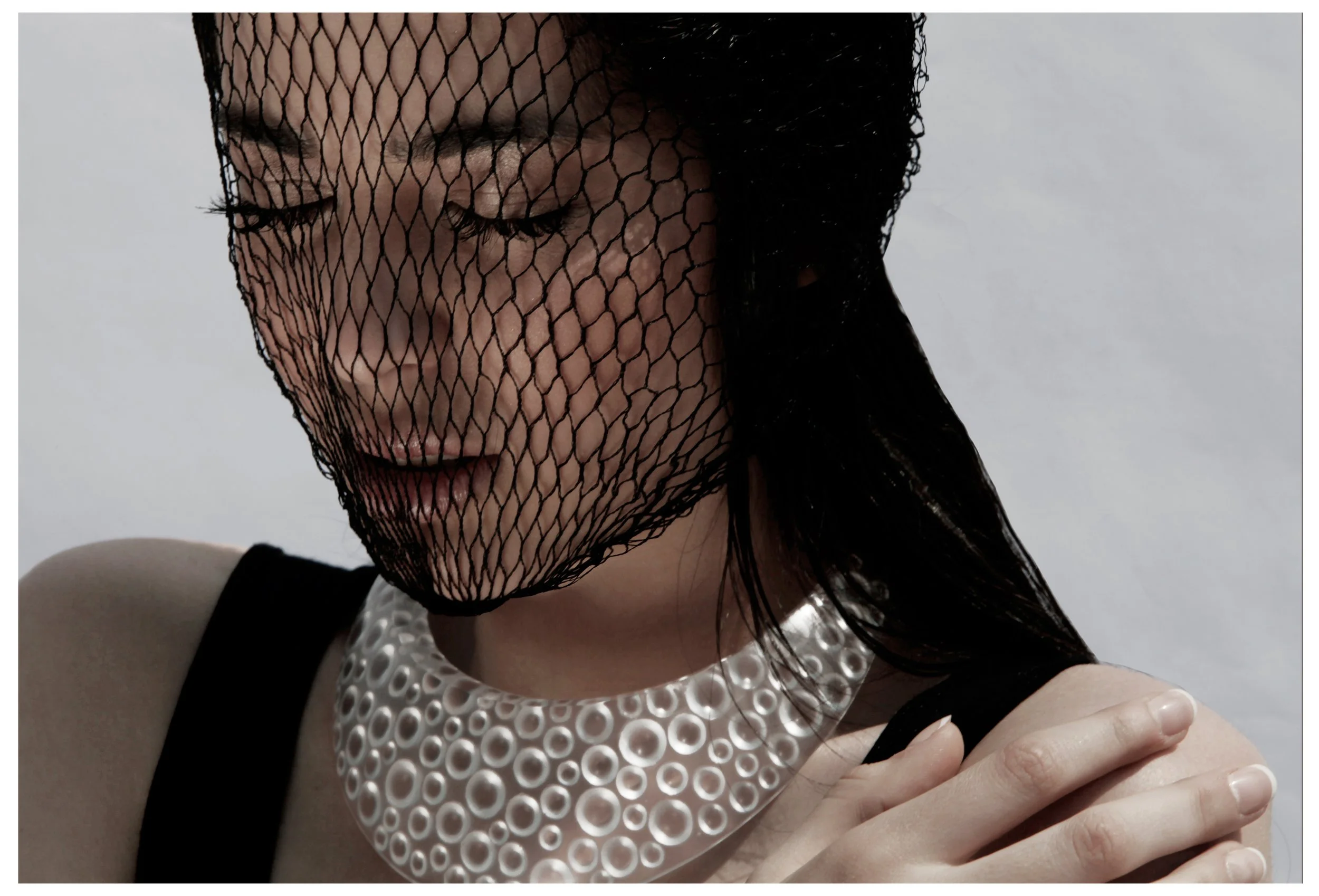 A woman with dark hair partially covered by a black fishnet veil, wearing a white textured outfit with black trim, touching her shoulder with her hand.
