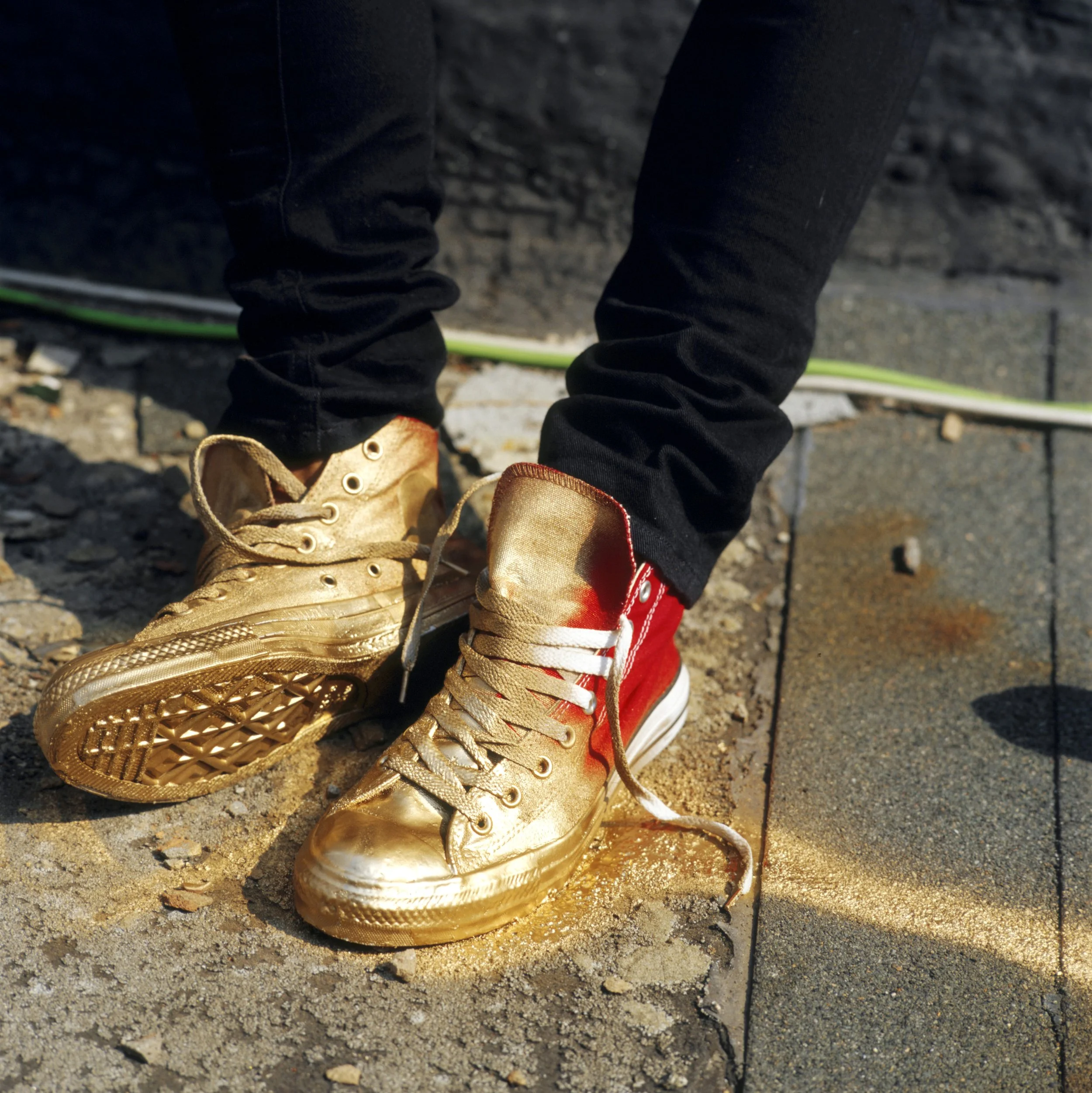 A pair of worn sneakers, one beige and the other red, on a gravel ground with a green cable and rocks nearby.
