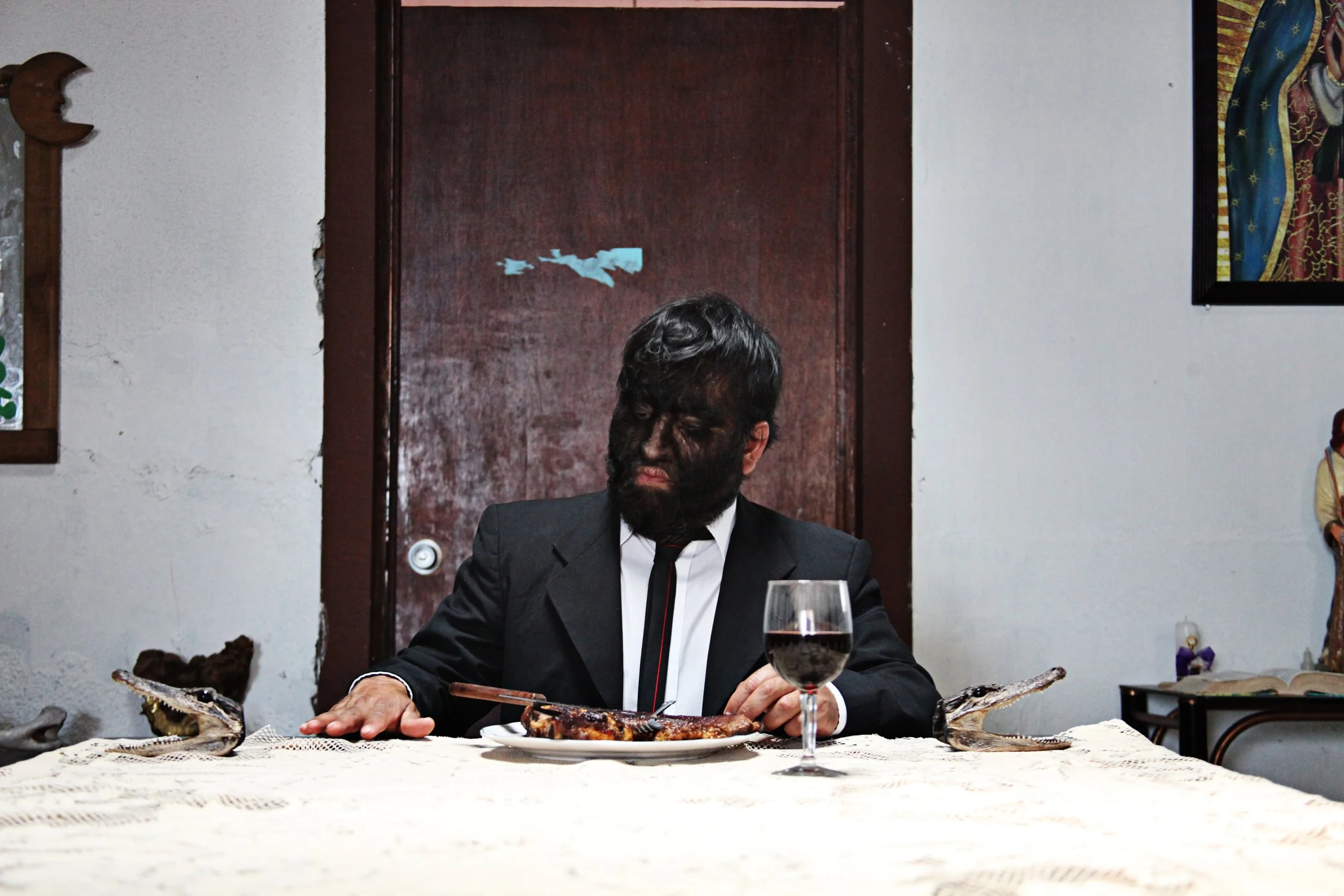 A man in a black suit and white shirt sitting at a table with a white lace tablecloth, dining on a large cooked fish with side dishes. There is a glass of red wine in front of him and an ornate fish-shaped serving dish on the table. The background fe