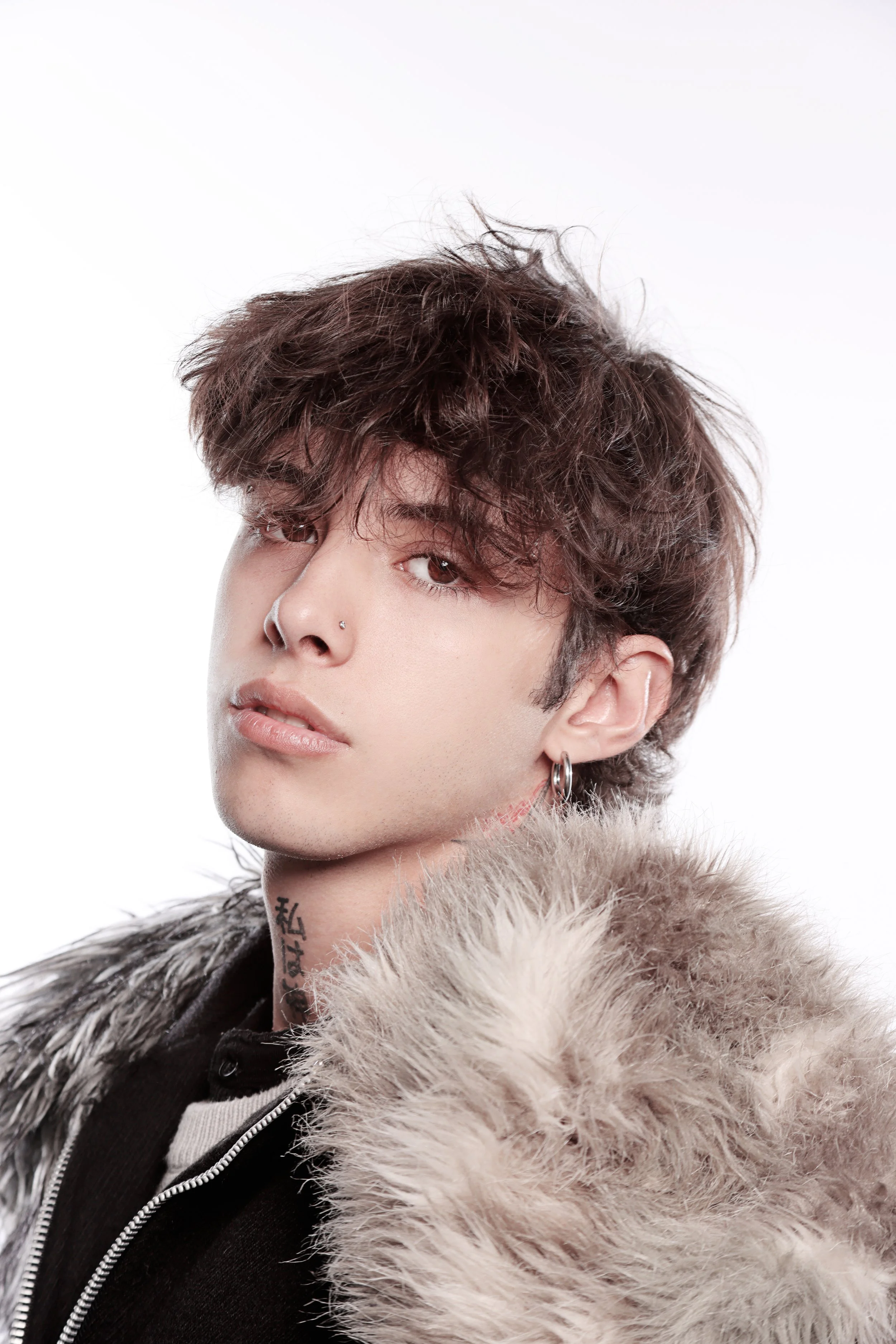 Close-up of a young man with brown curly hair, piercings, tattoos, wearing a fur-lined coat against a plain white background.