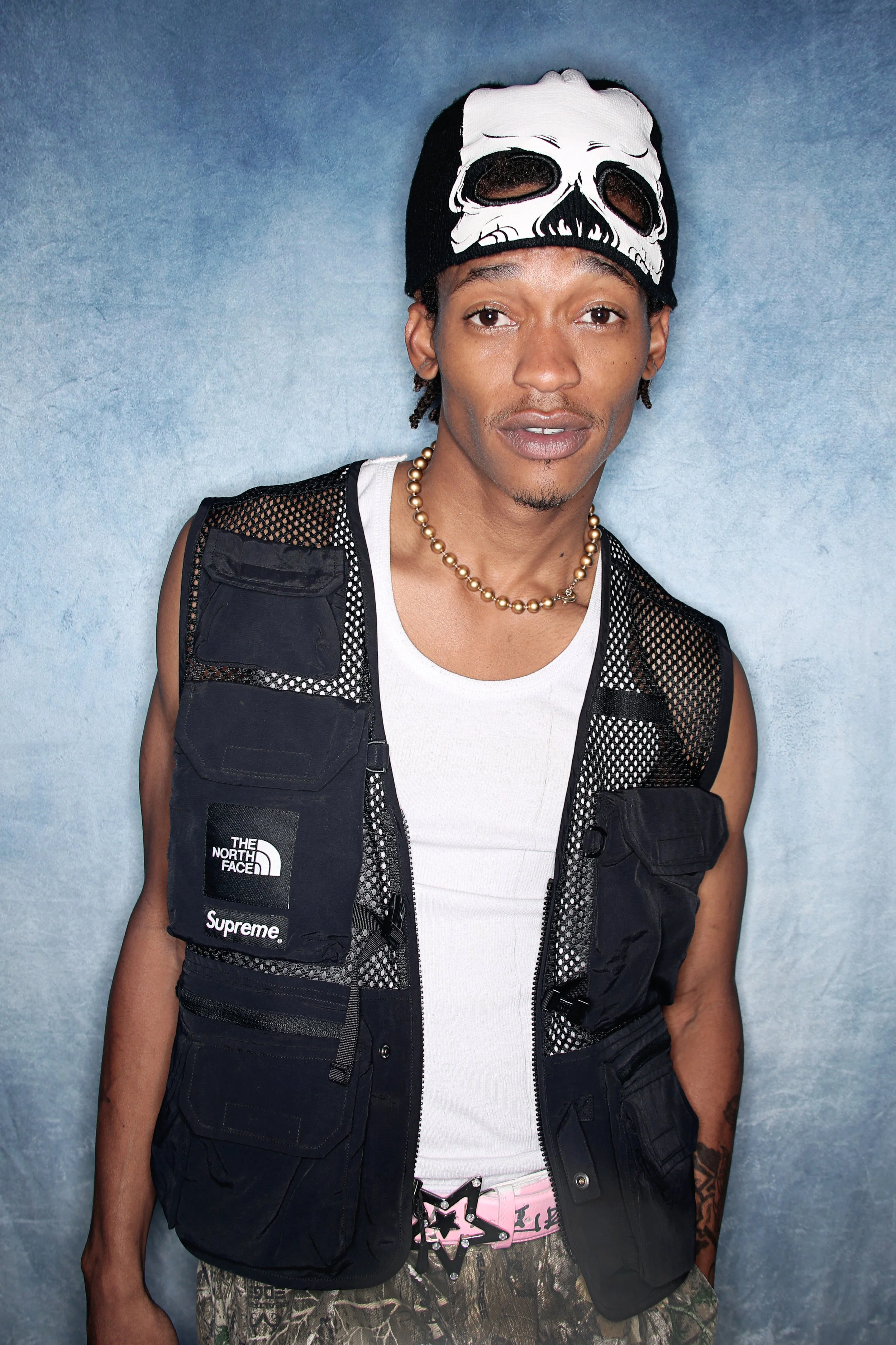 A young man wearing a black skull mask on his forehead, a black vest with multiple pockets and logos, a white tank top, camouflage pants, a pink belt with a star-shaped buckle, and gold necklace, standing against a blue background.