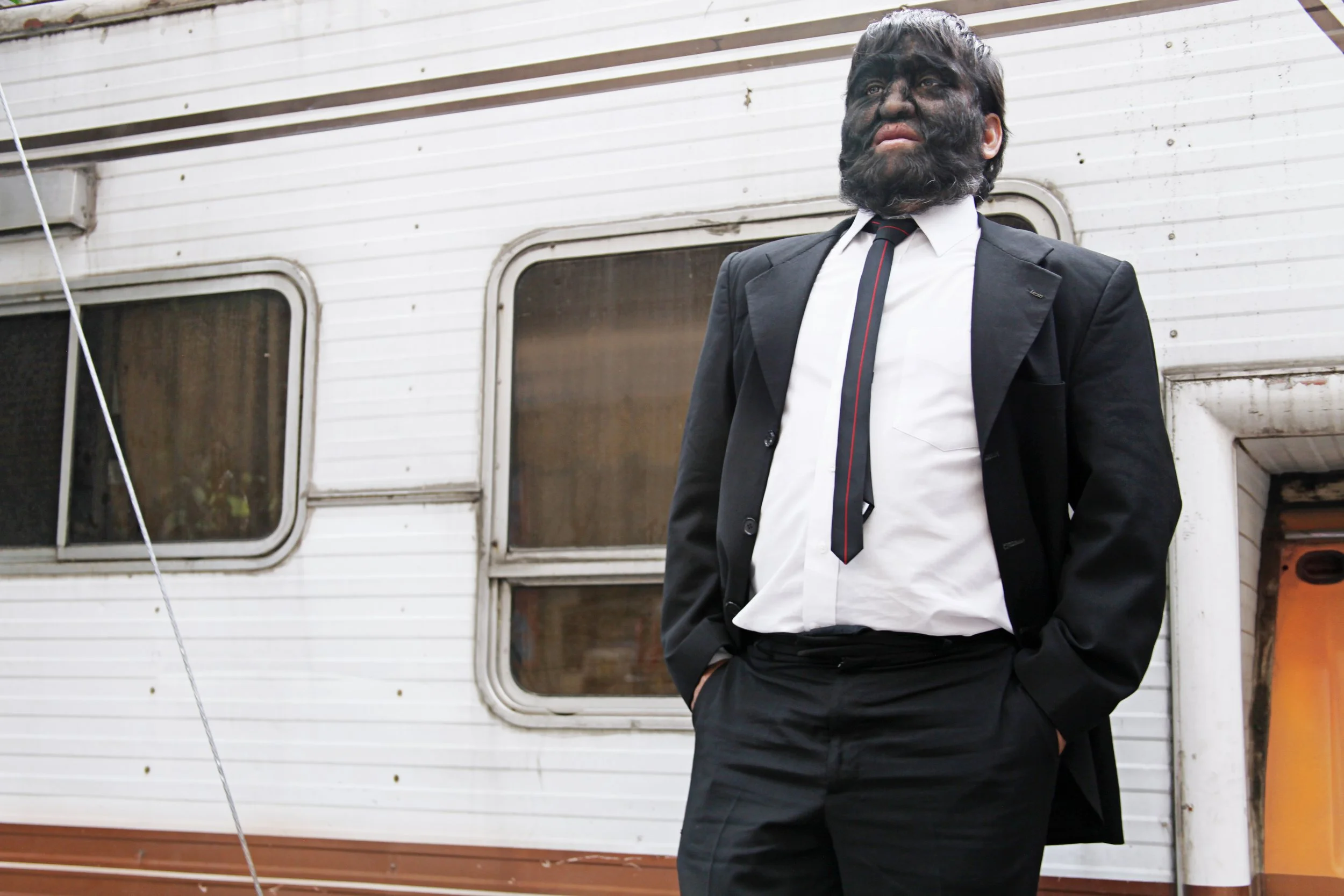 A man in a black suit, white shirt, and black tie standing outdoors in front of an old, weathered camper trailer, with his hands in his pockets.