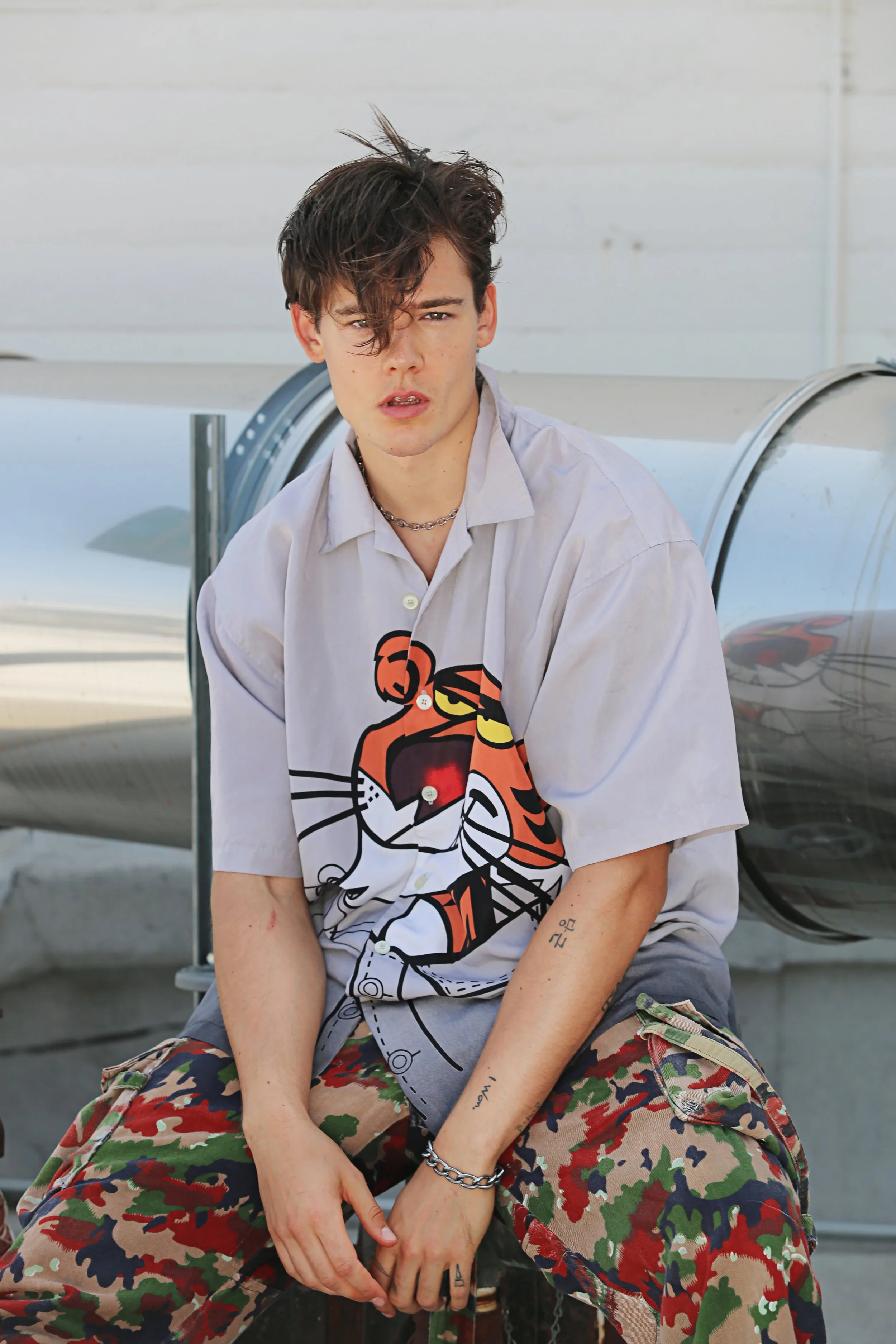 A young man with dark, messy hair, sitting outdoors near a reflective surface, wearing a graphic shirt with a tiger face, camouflage pants, and jewelry, looking directly at the camera with a serious expression.