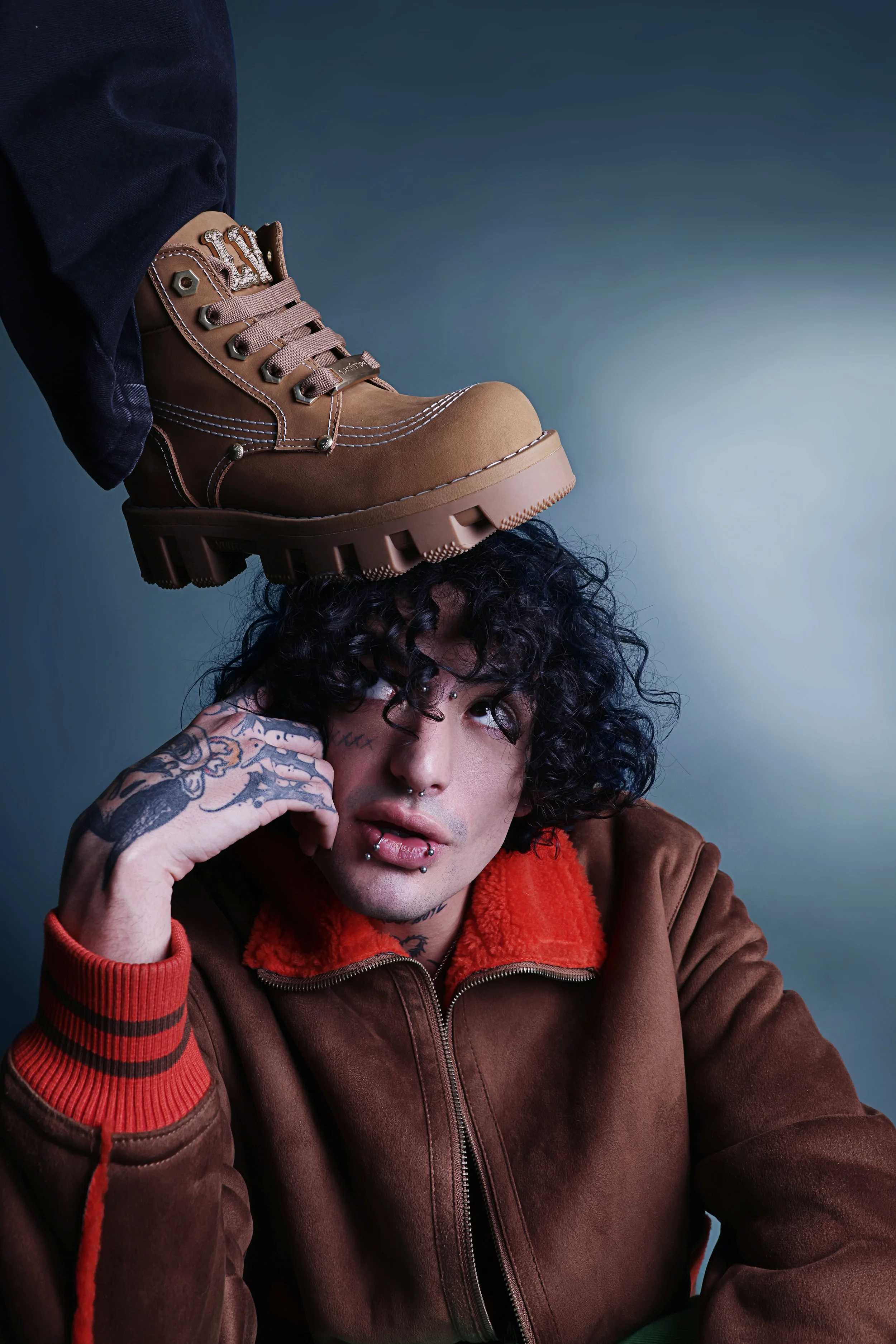 A person with tattoos on their hand and face, wearing a brown jacket with red accents, resting their head on their hand, and looking directly at the camera.