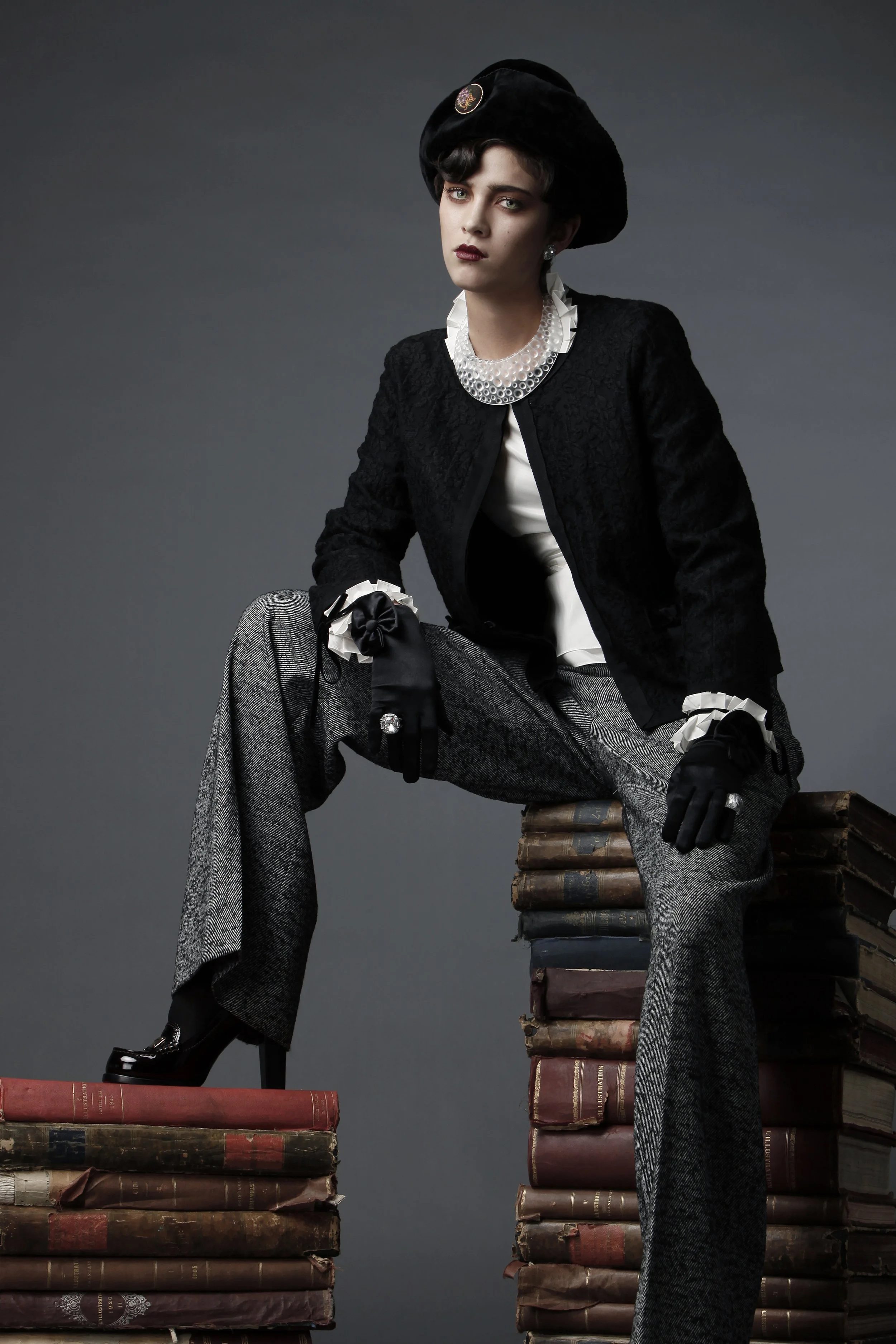 A woman dressed in vintage clothing with a black hat, pearl necklace, and lace jacket sits on a stack of old books against a dark background.