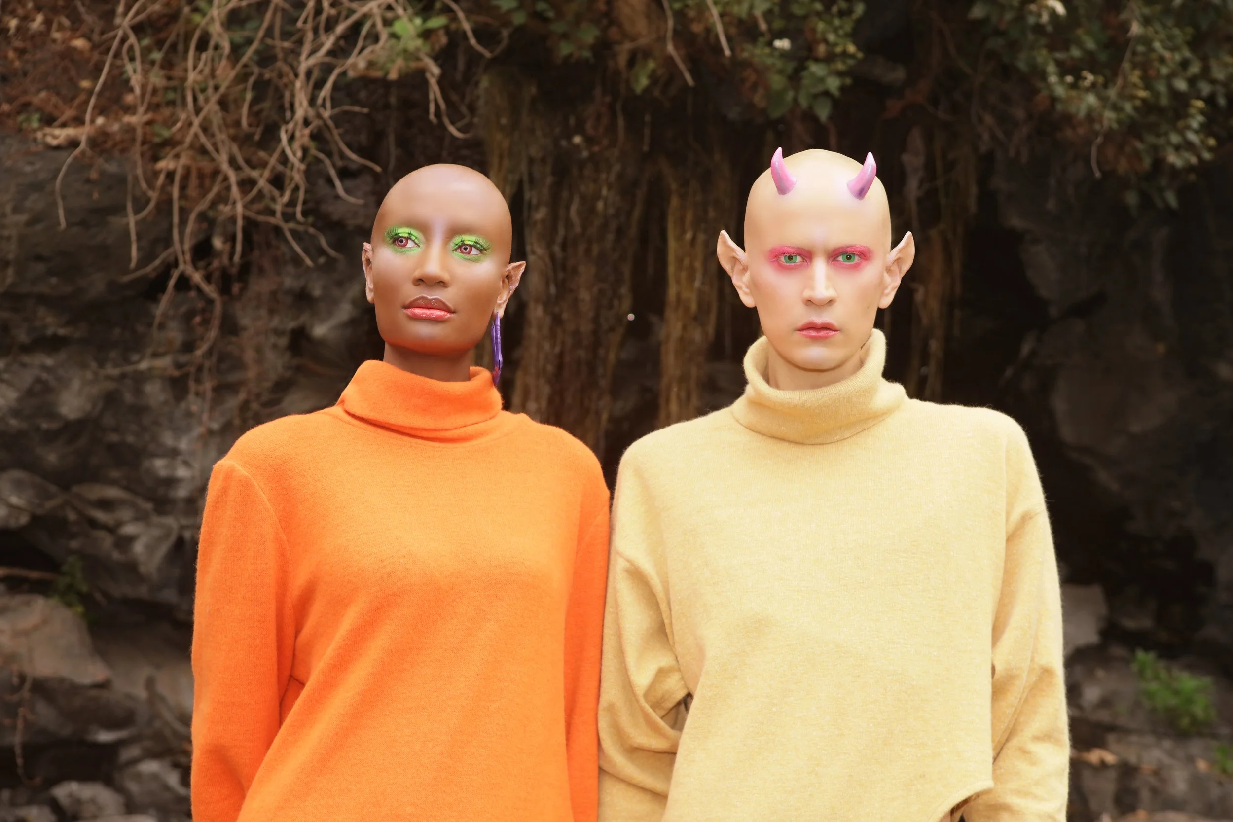Two individuals with bald heads wearing brightly-colored outfits, standing outdoors with a rocky, earthy background. The person on the left has green makeup around the eyes and a purple earring, wearing an orange turtleneck sweater. The person on the