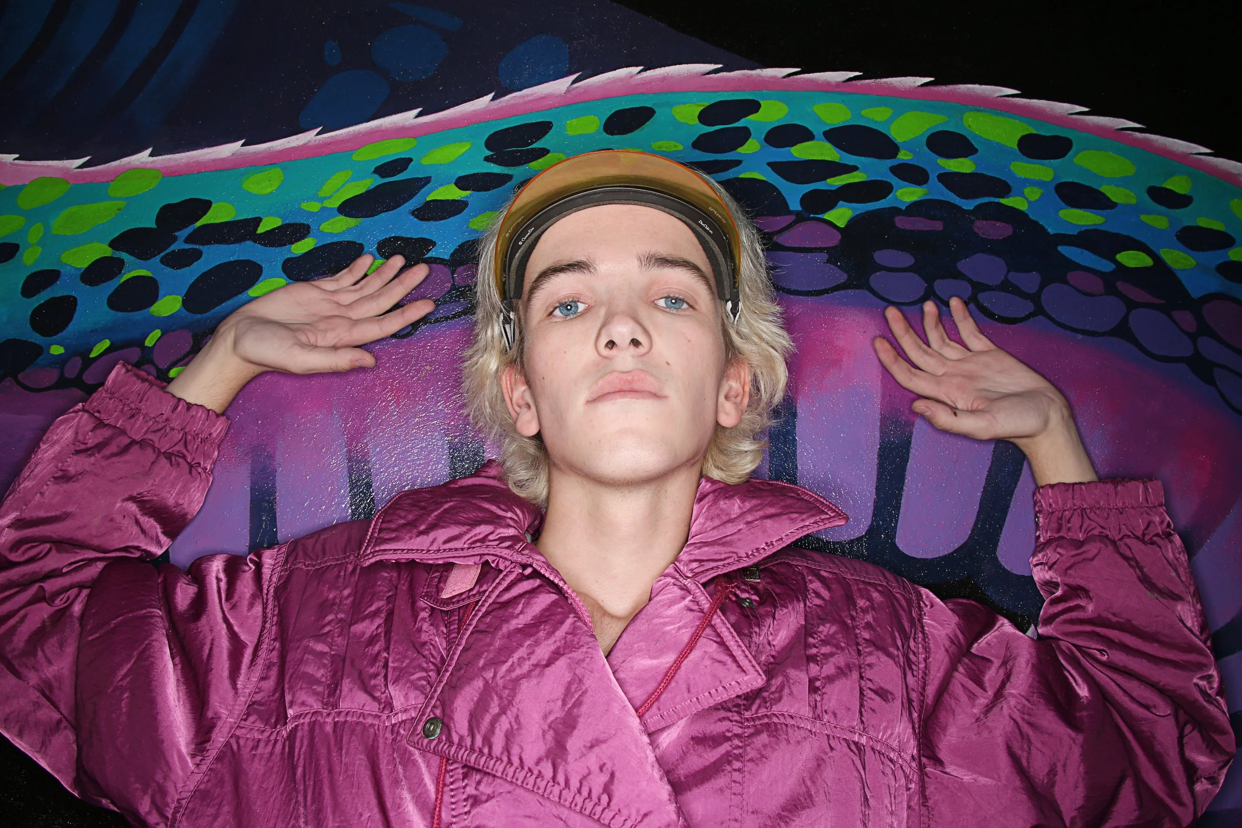 Person with blonde hair, wearing a pink jacket, lying on a colorful abstract painted surface with their hands behind their head.