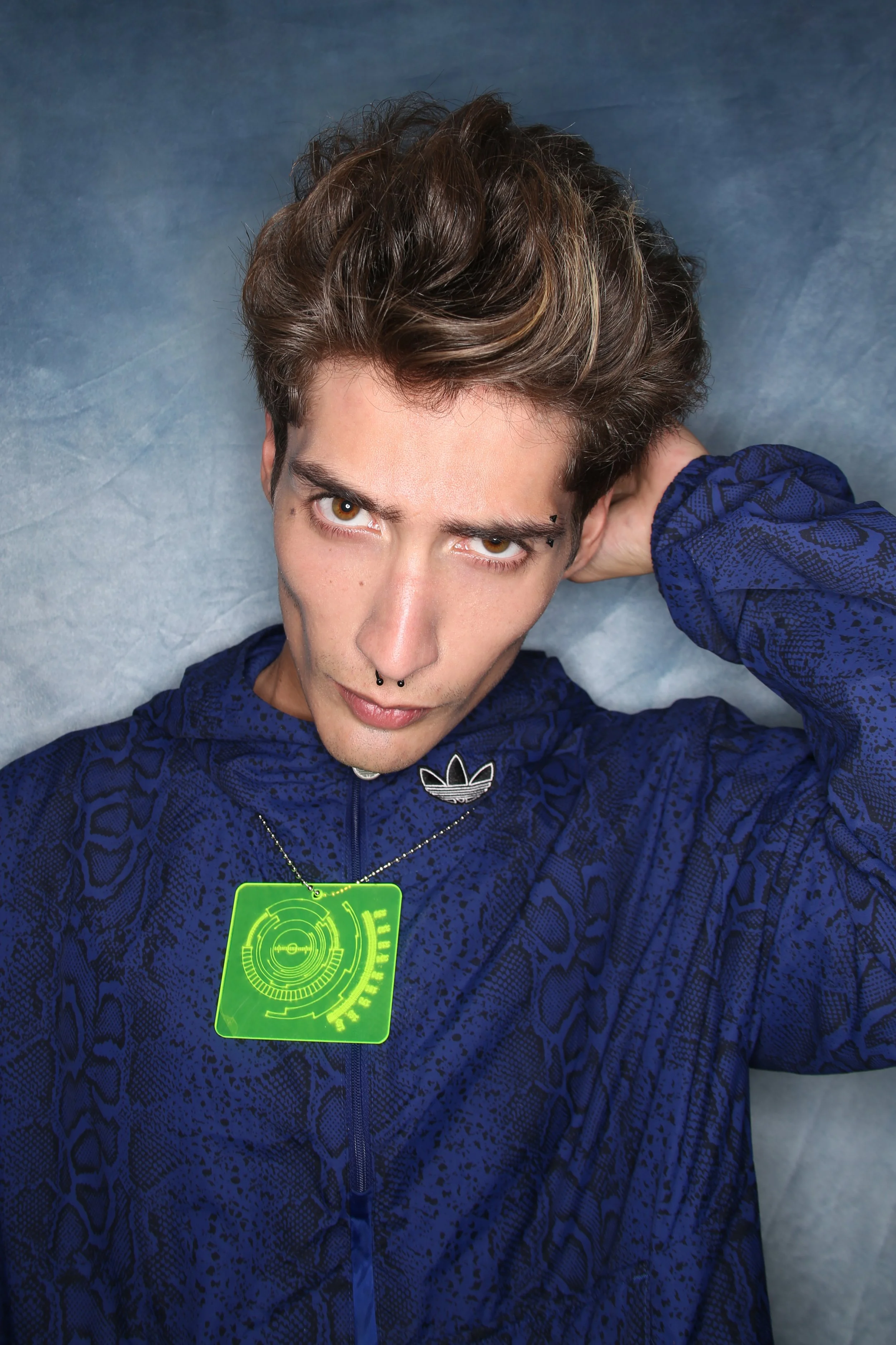 Young man with brown hair, light skin, hazel eyes, and a septum piercing, wearing a blue patterned jacket and a neon green digital badge, lying on a blue surface and looking into the camera.