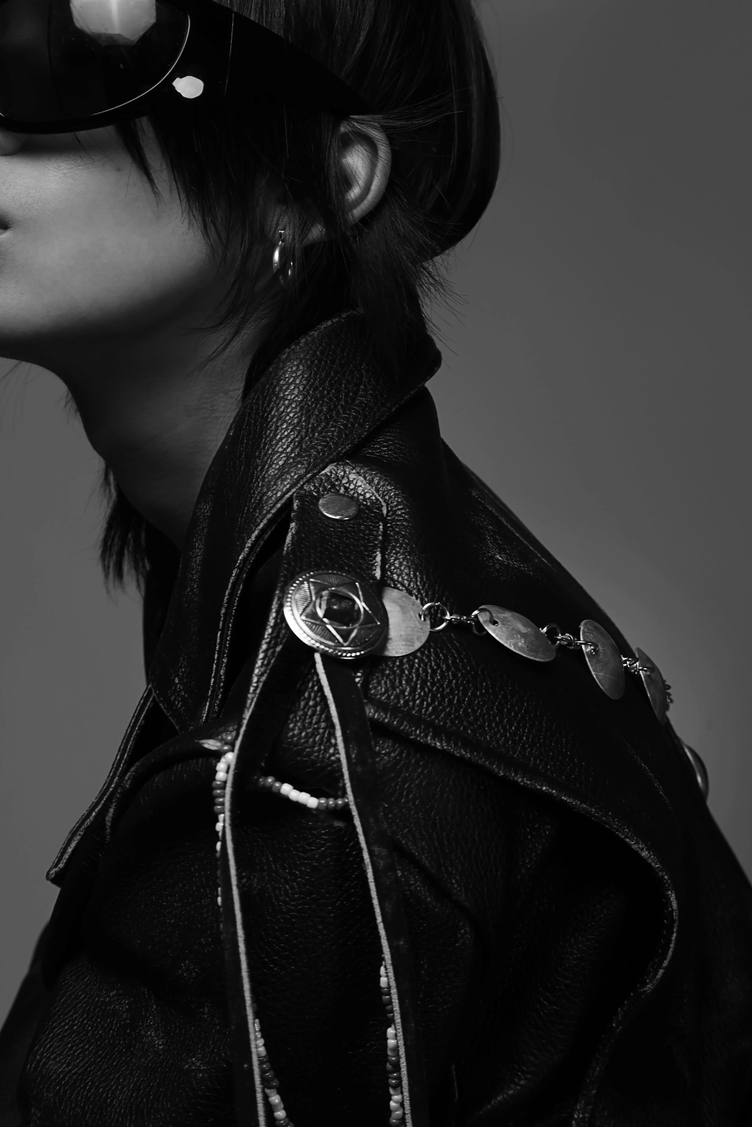 Close-up black and white photograph of a woman wearing sunglasses, a earring, a leather jacket with pins, and layered necklaces.