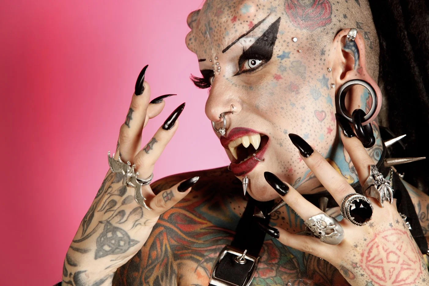 A person with extensive tattoos, piercings, and gothic makeup, posing with their teeth showing and fingers near their face, with a pink background.