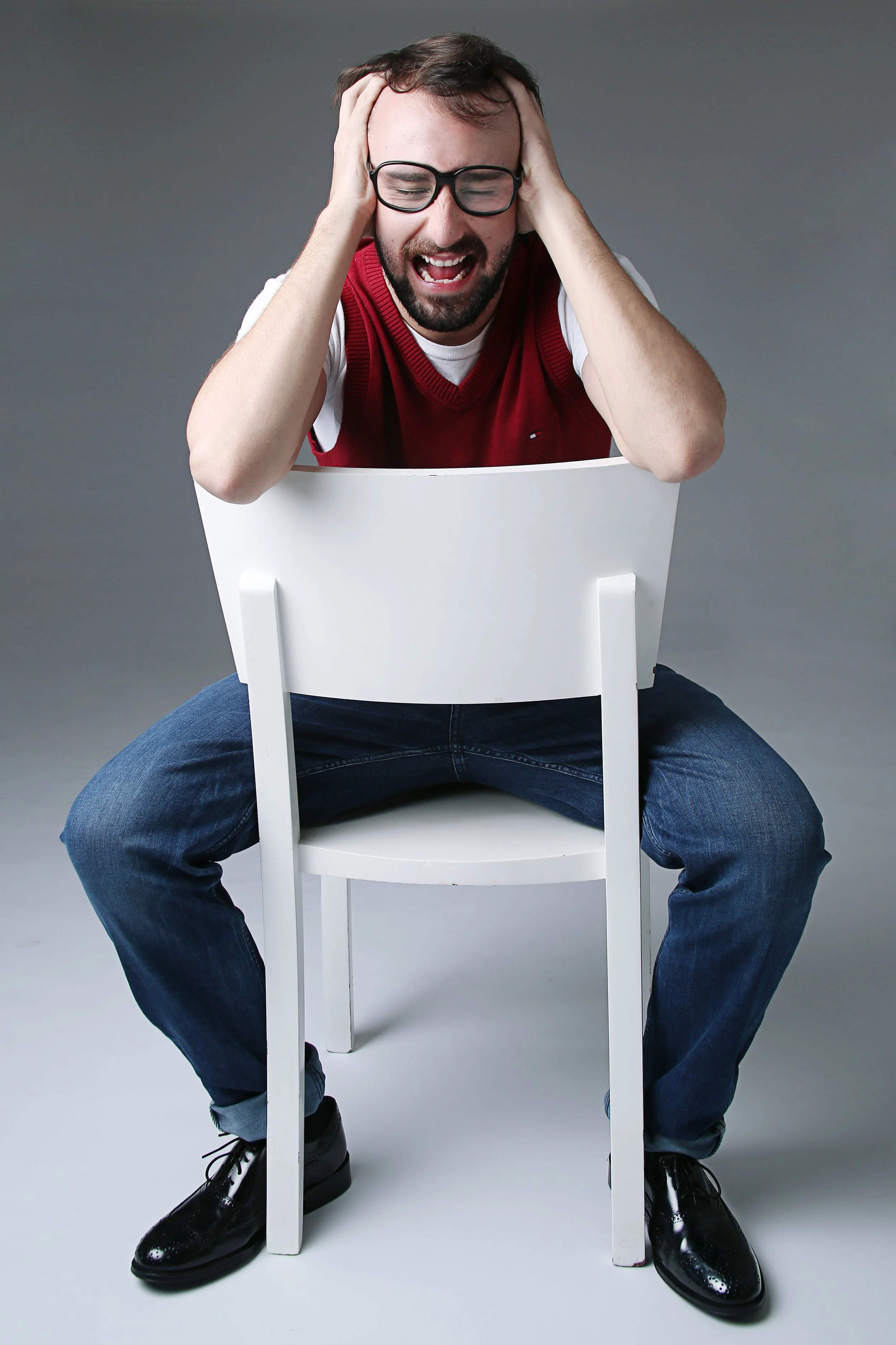 A man wearing glasses and a red vest, sitting on the floor with a white chair in front of him, holding his head with both hands, appearing distressed or overwhelmed.
