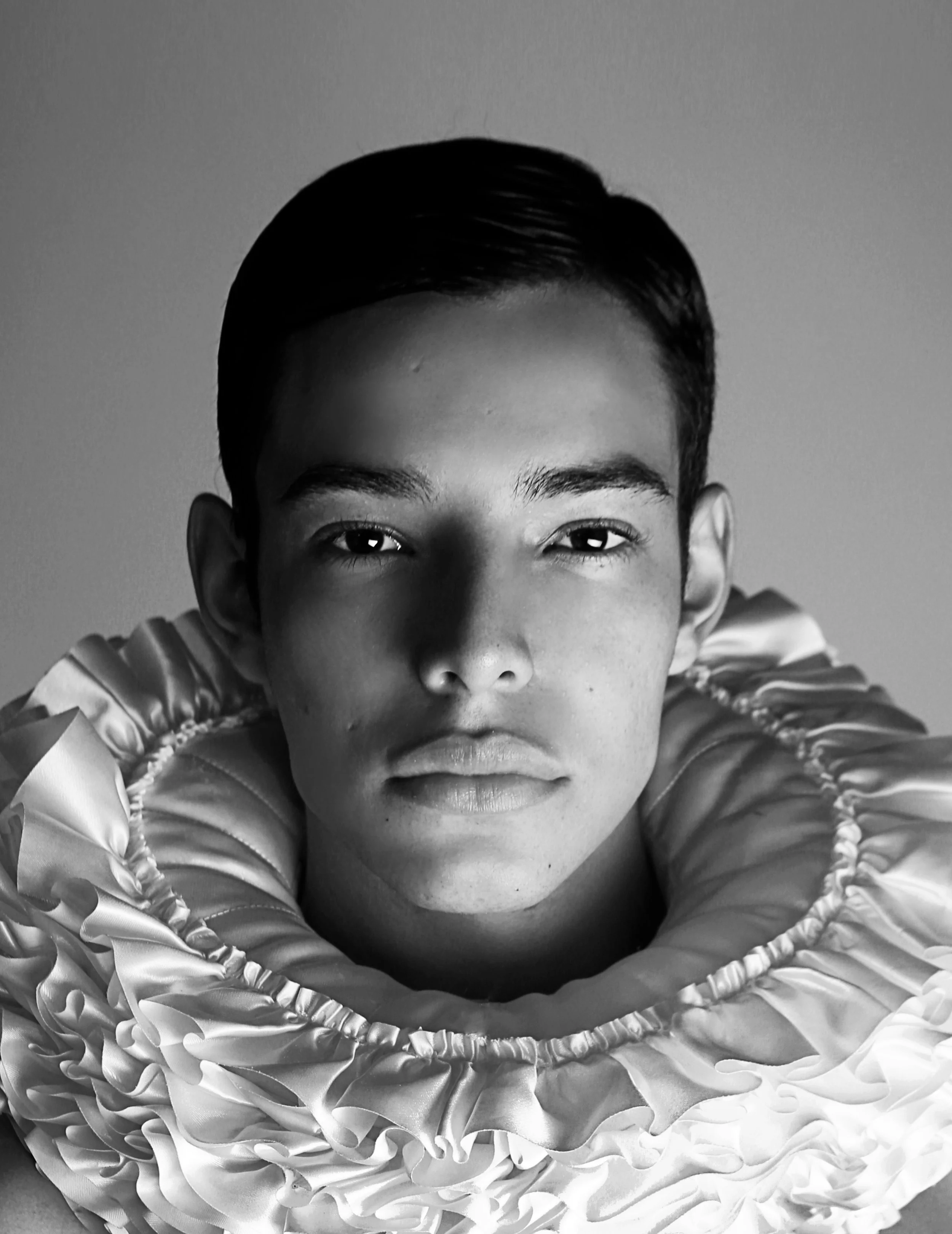 A young boy with dark hair and a focused expression lying down with his face close to the camera, surrounded by ruffled fabric or material.