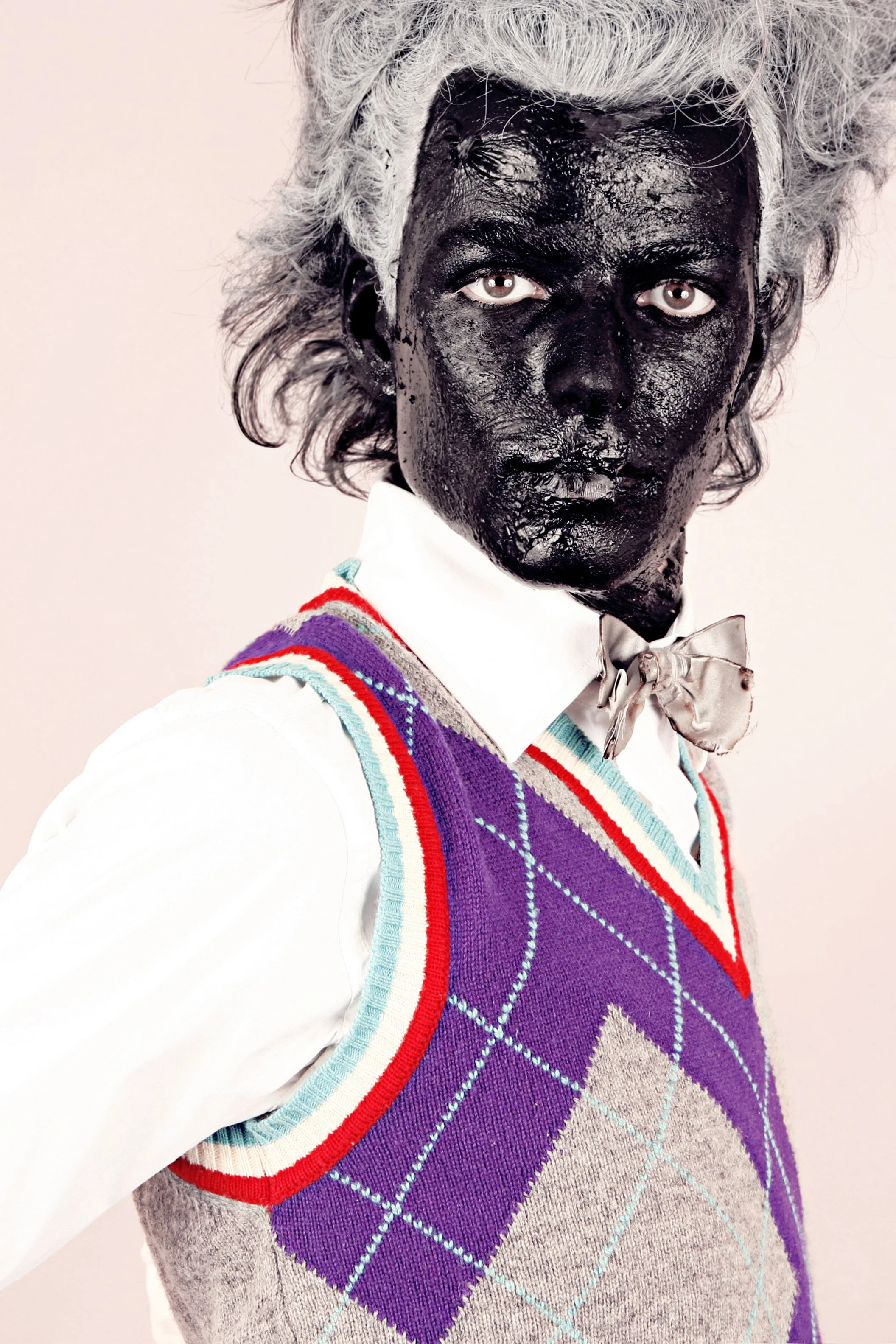 Person with a black painted face and white hair, wearing a white shirt, a beige bow tie, and a colorful argyle vest.