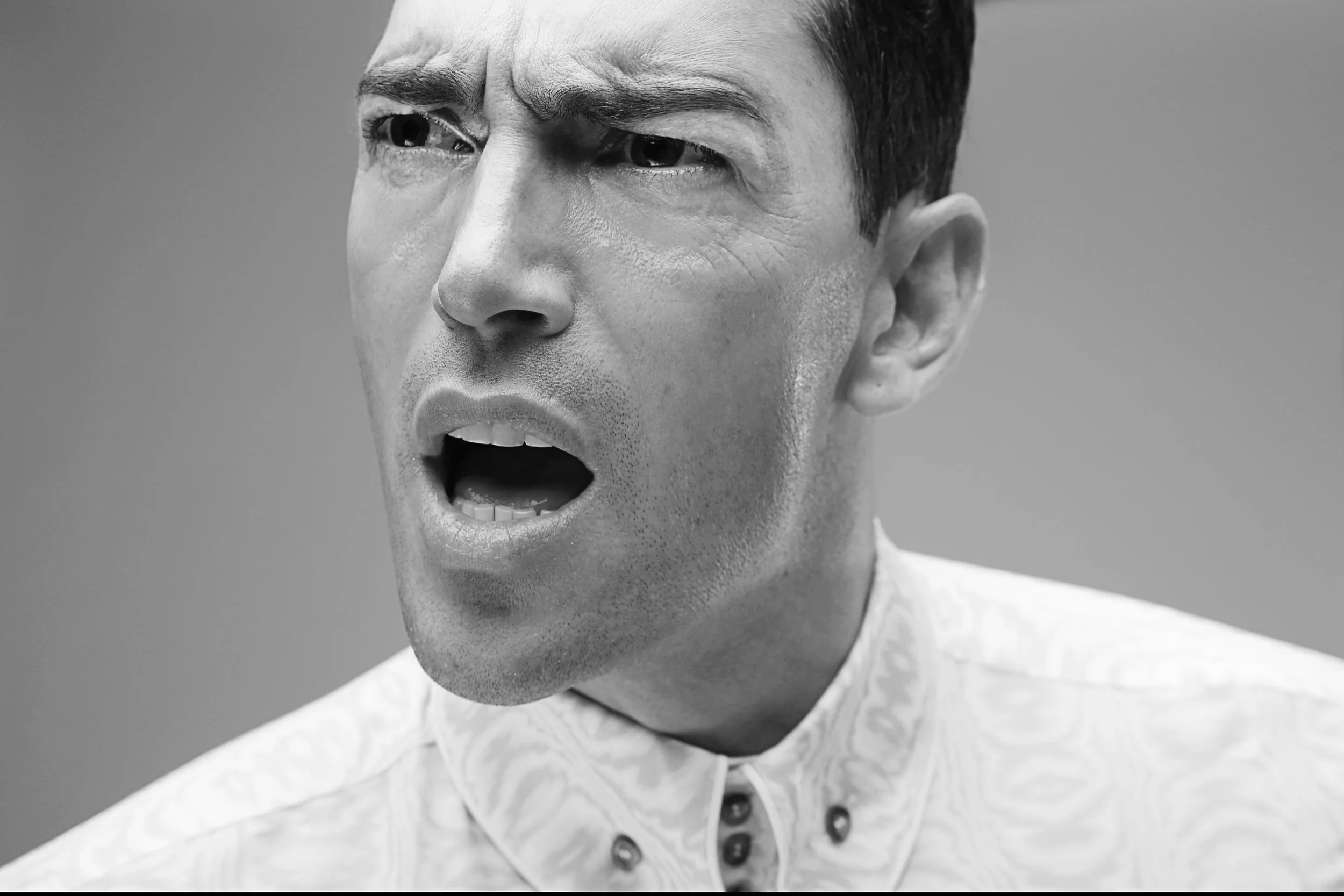 Close-up of a man's face with one eye squinting and his mouth open, expressing shock or surprise, in black and white.